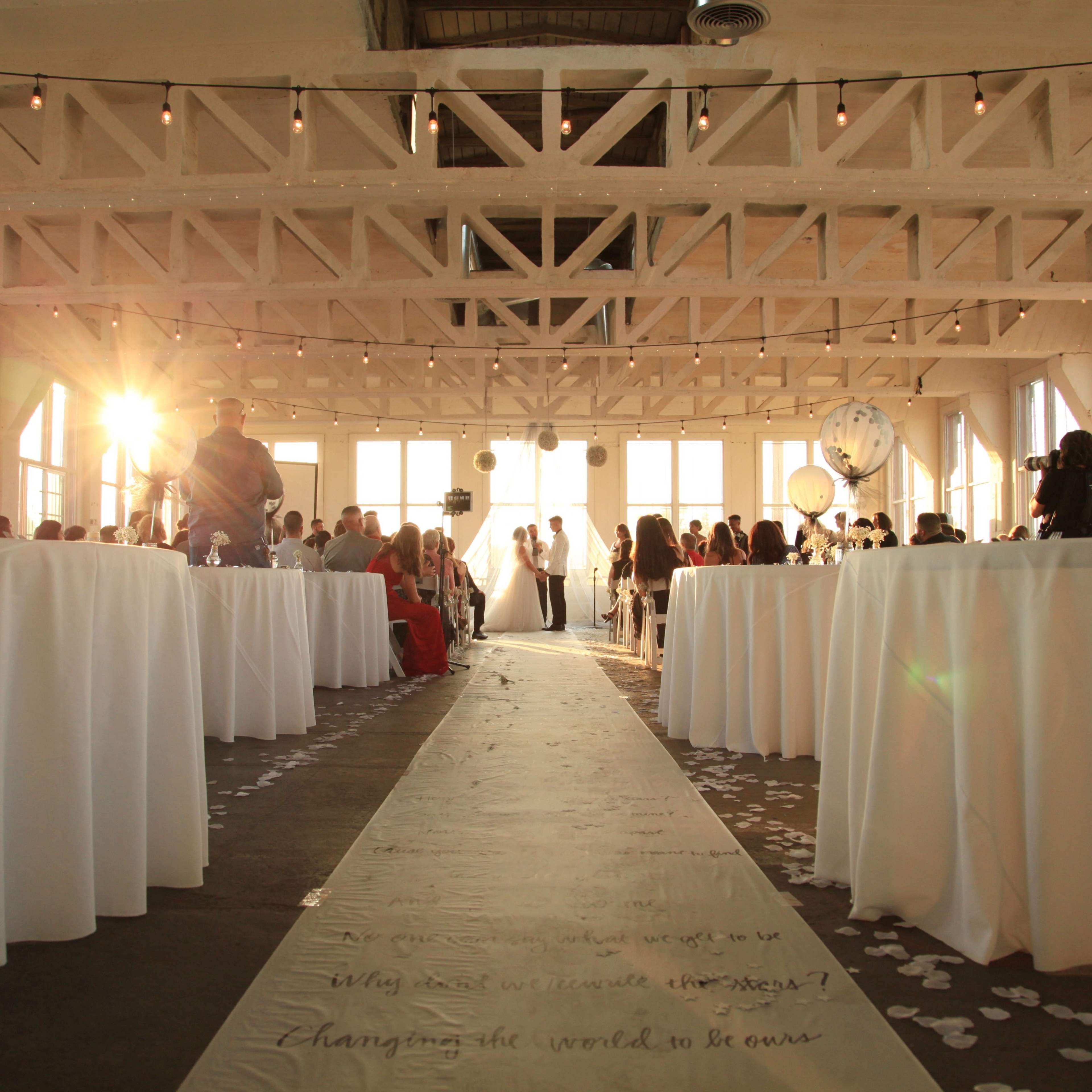 A couple exchanges vows in a well-decorated indoor venue with large windows, surrounded by seated guests and elegant table settings.