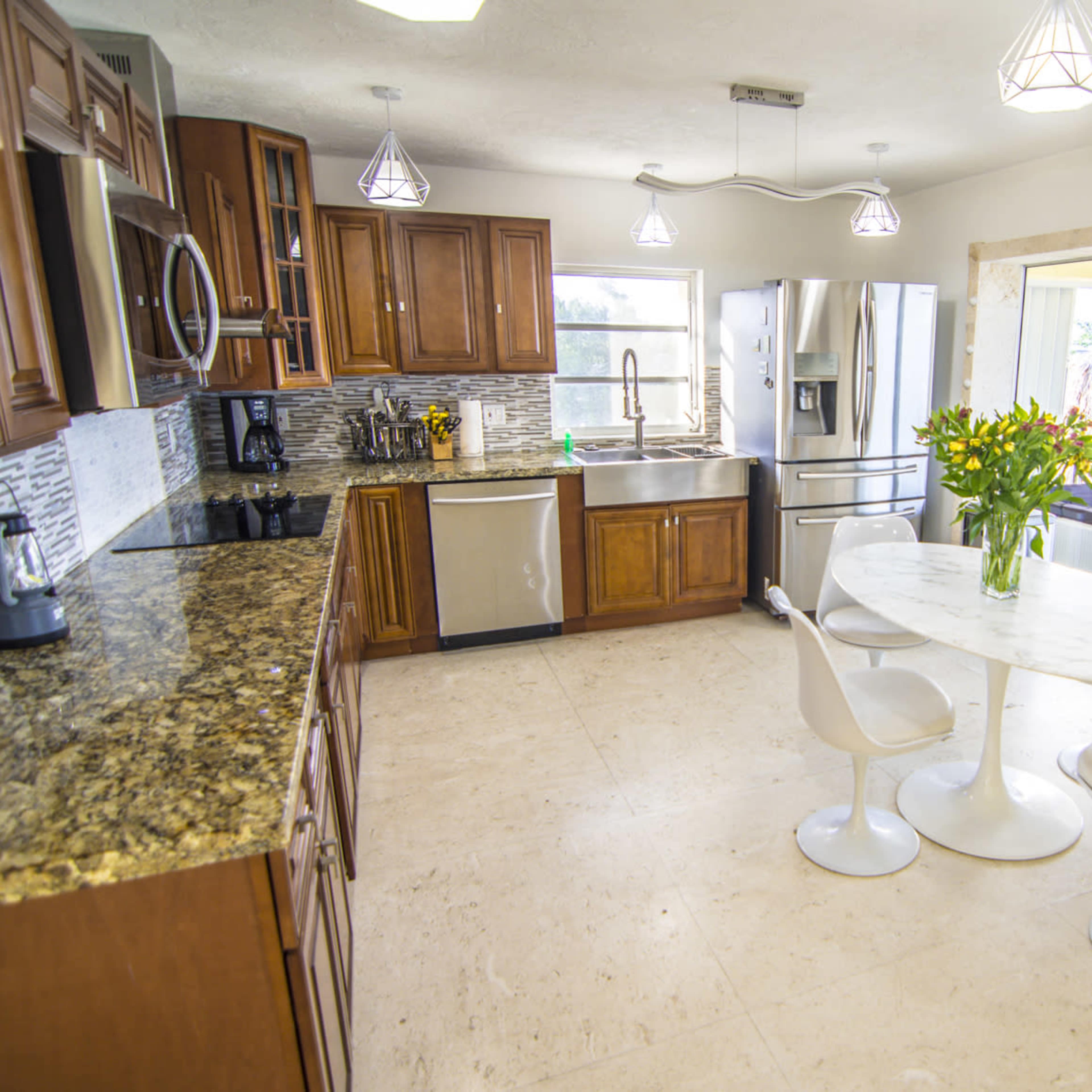 A modern kitchen features wooden cabinets, stainless steel appliances, a marble table with three chairs, and large windows allowing natural light.