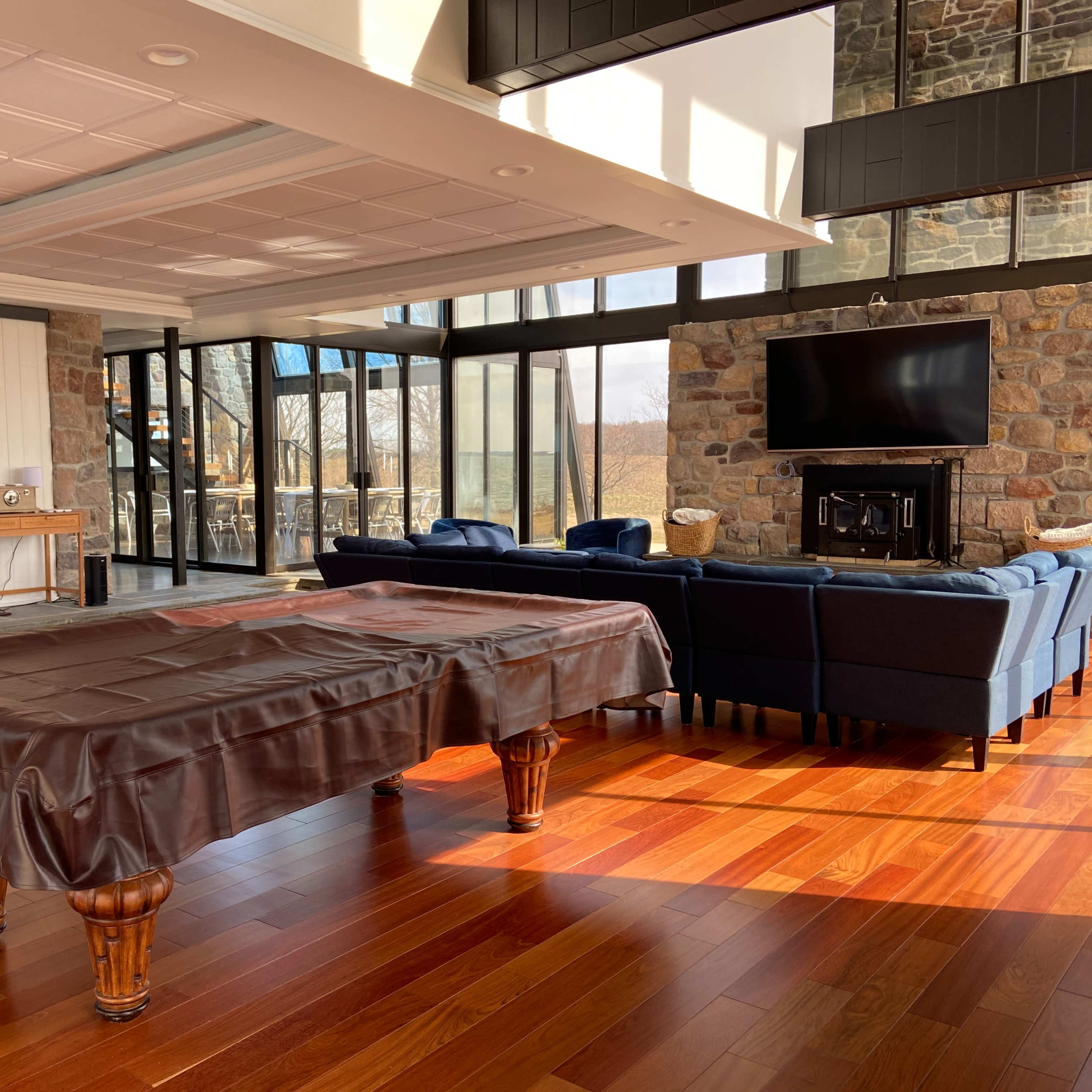 A modern indoor space featuring a pool table covered with a brown cloth, surrounded by large windows, a stone fireplace, and a seating area with blue sofas.