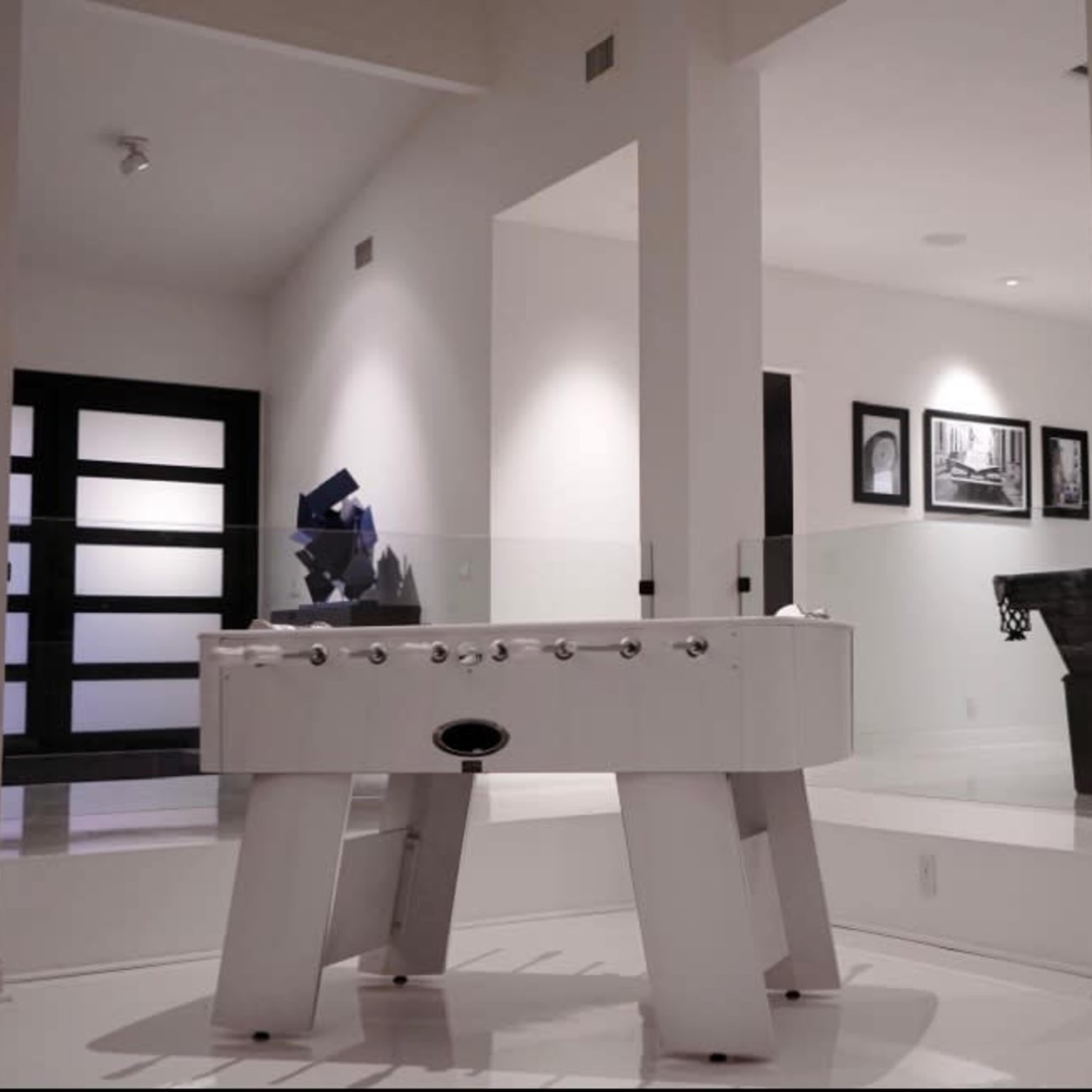 A modern room featuring a white foosball table in the foreground and a black piano in the background, with large windows and gallery-style artwork on the walls.