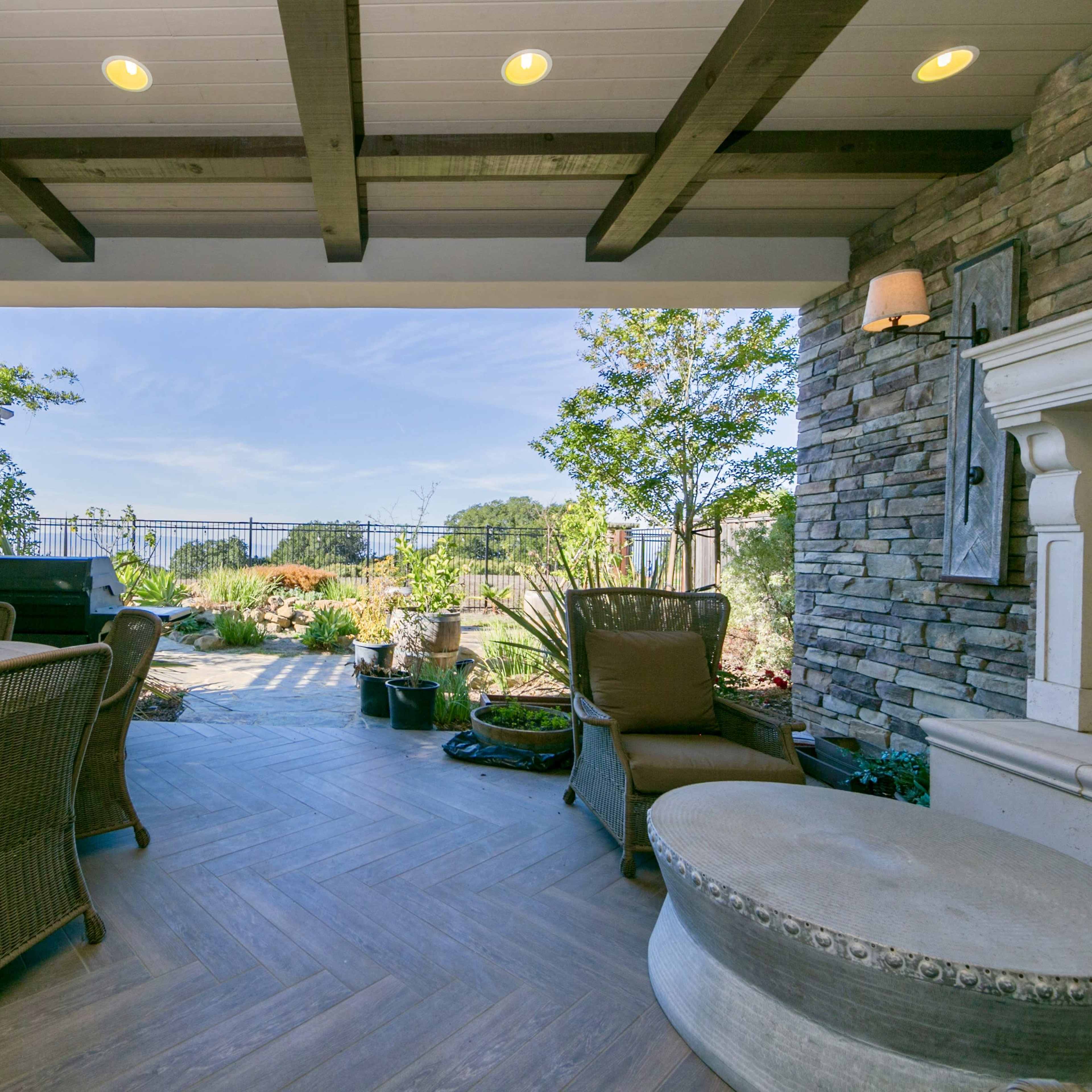 A covered outdoor patio with a wooden table and wicker chairs, overlooking a landscaped garden.