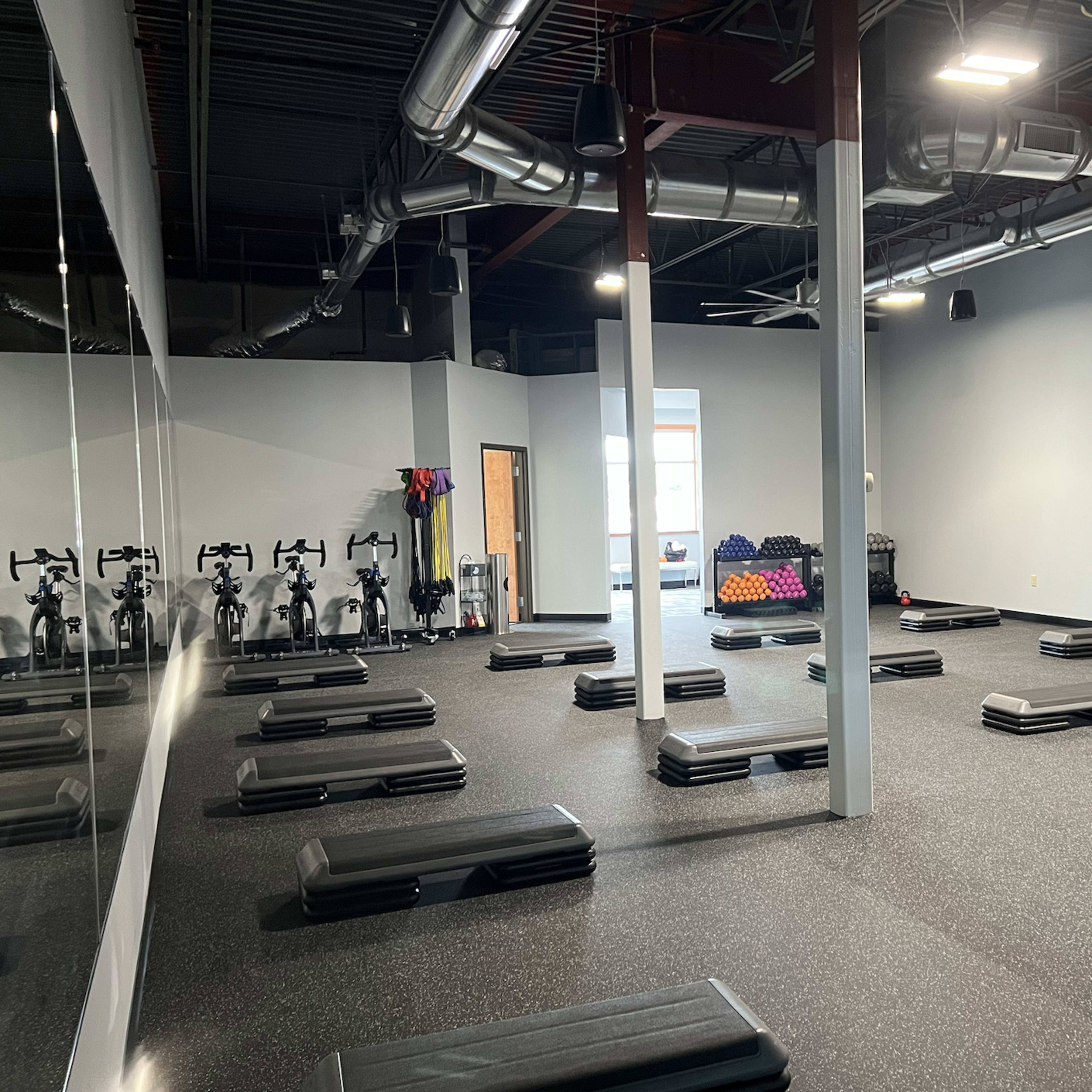 A spacious fitness studio with numerous step platforms arranged on the black rubber floor, exercise bikes lined up against the wall, and a few weights scattered in one corner.