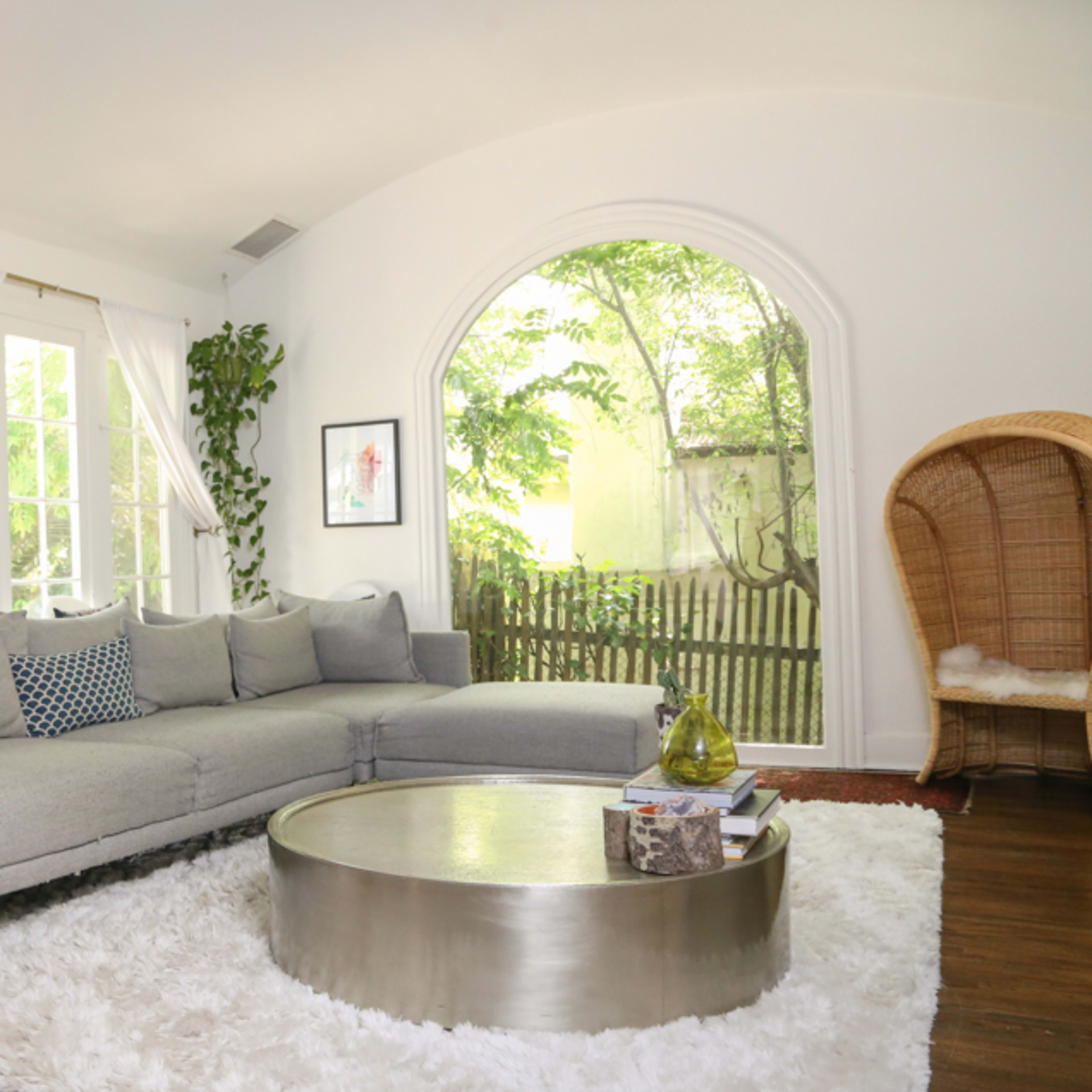 A cozy living room with a gray sectional sofa, a round coffee table, and a large window overlooking a green outdoor space.