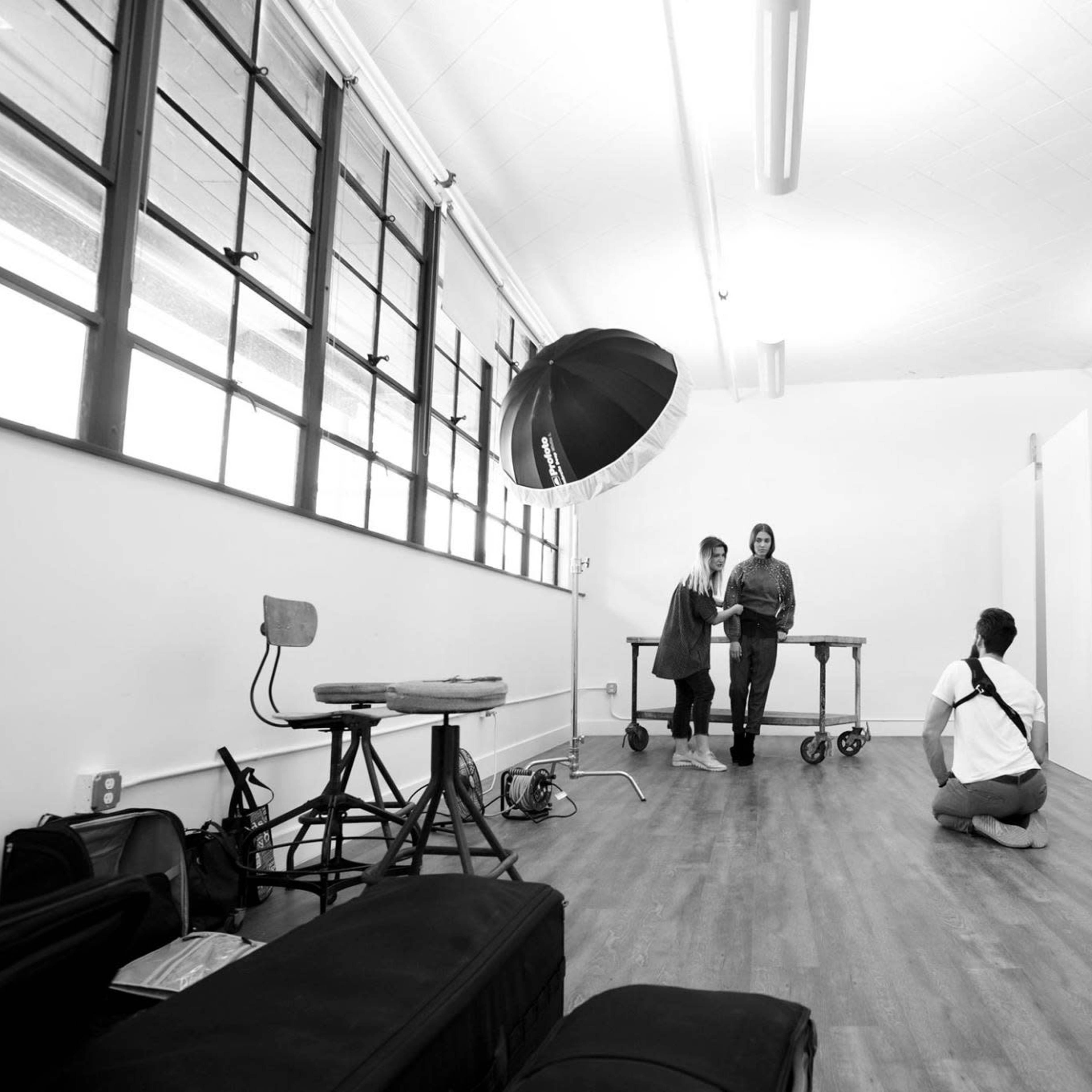 Two individuals stand beside a table in a spacious studio with large windows, while a third person crouches on the floor, preparing to take a photograph.