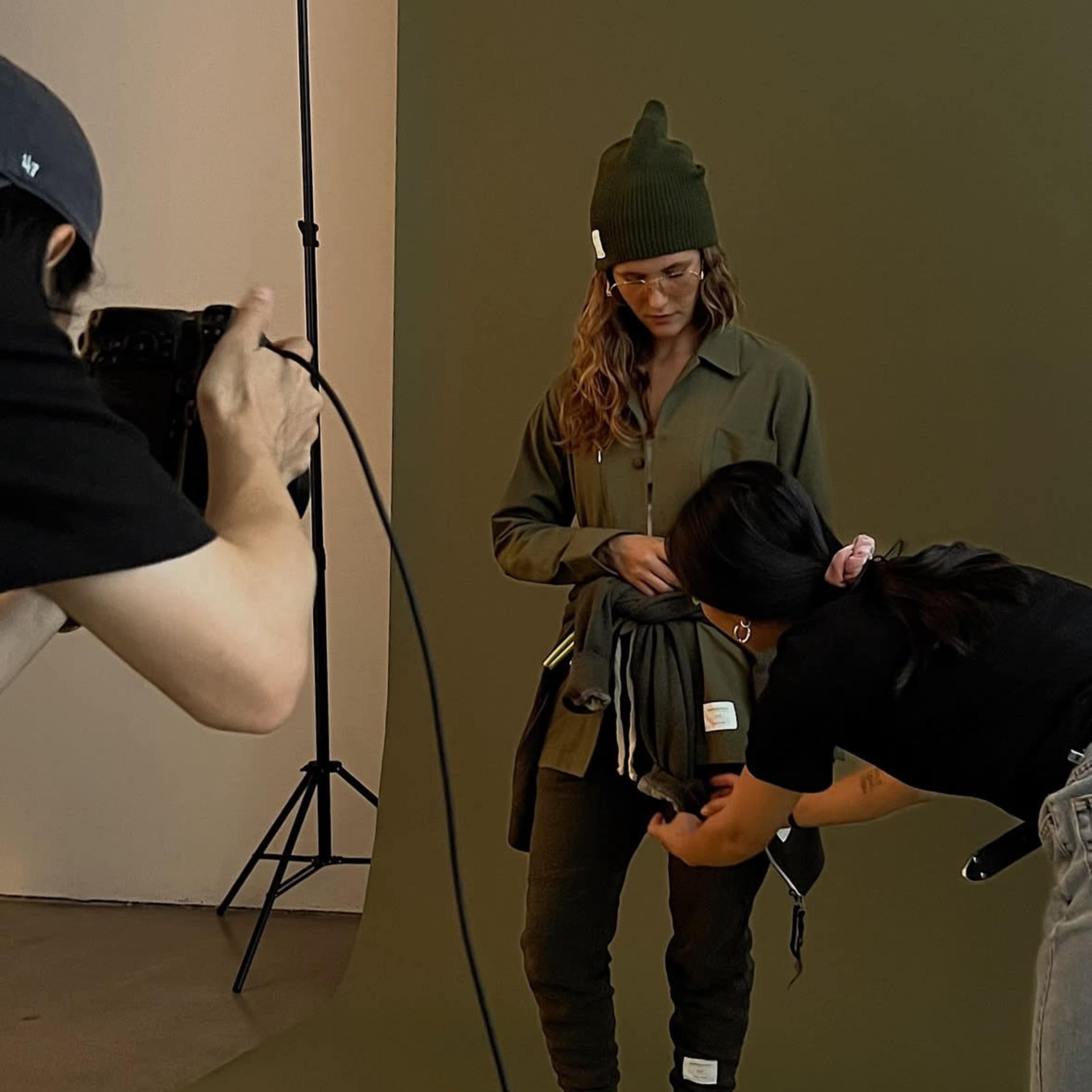 A model poses in a green outfit while a stylist adjusts her clothing in a studio setting with a neutral backdrop.
