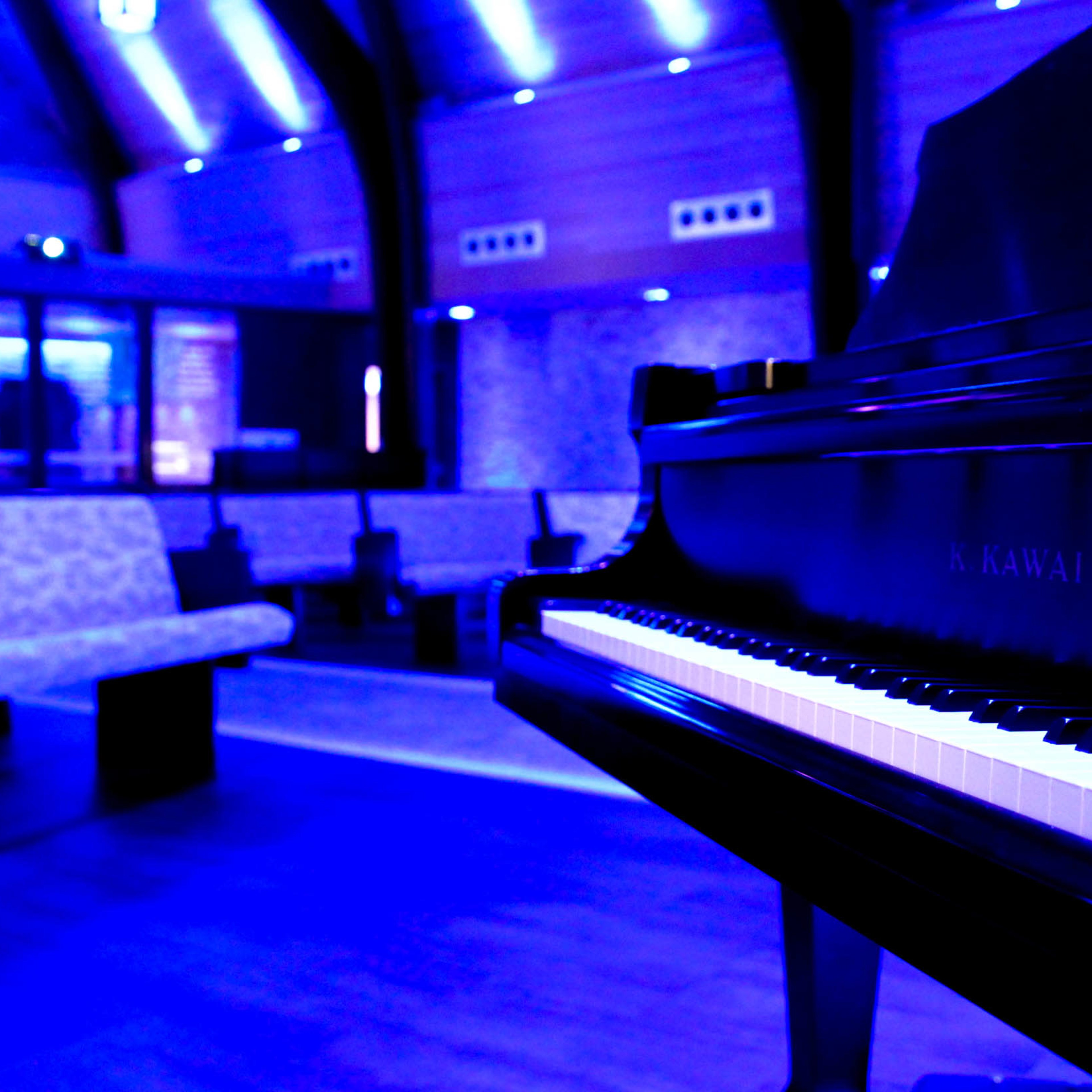 A black grand piano is positioned in a modern room with blue lighting and minimalist seating.