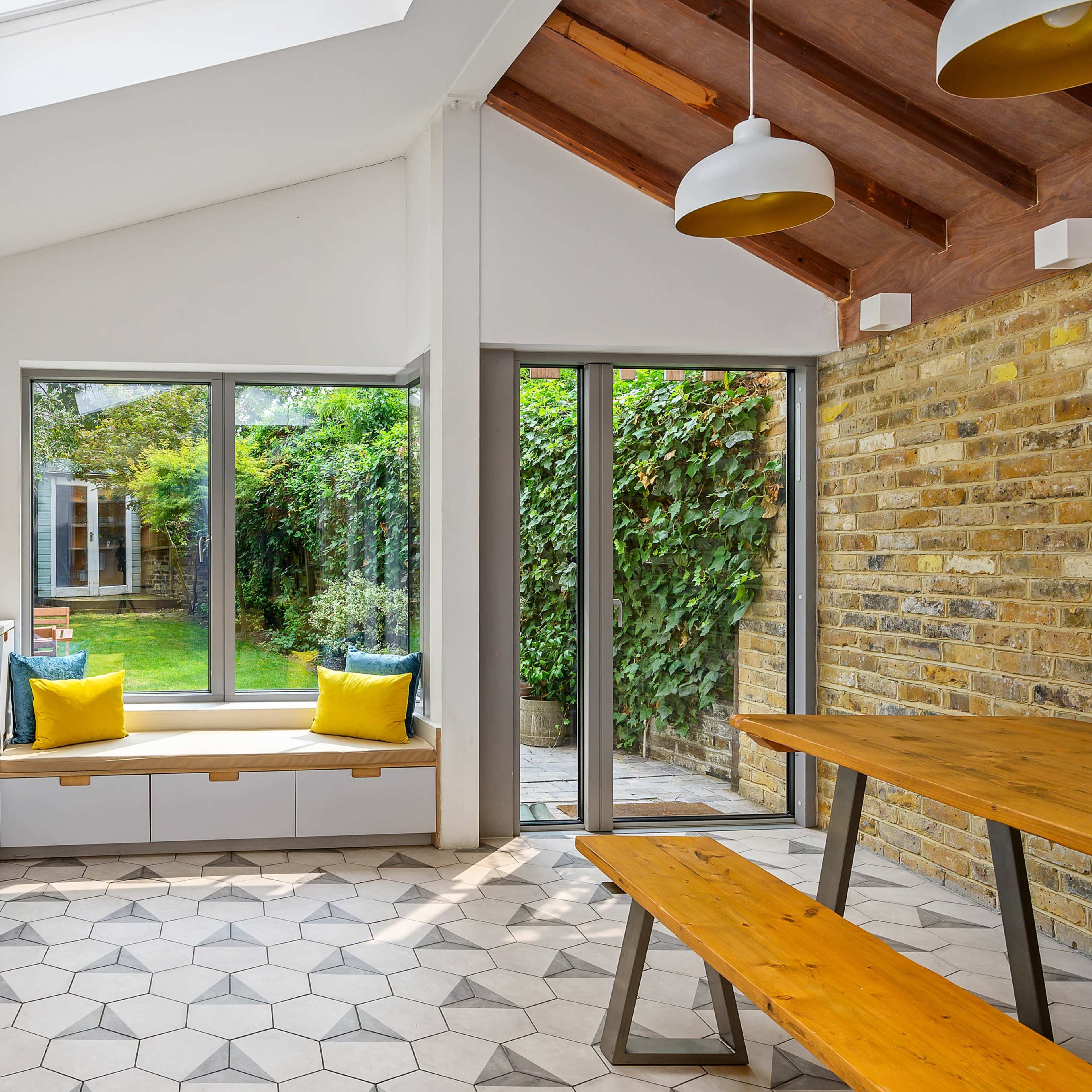 A modern kitchen with a skylight, featuring white cabinetry, a yellow tiled backsplash, and a dining area with a wooden table and bench seating by large windows overlooking a garden.
