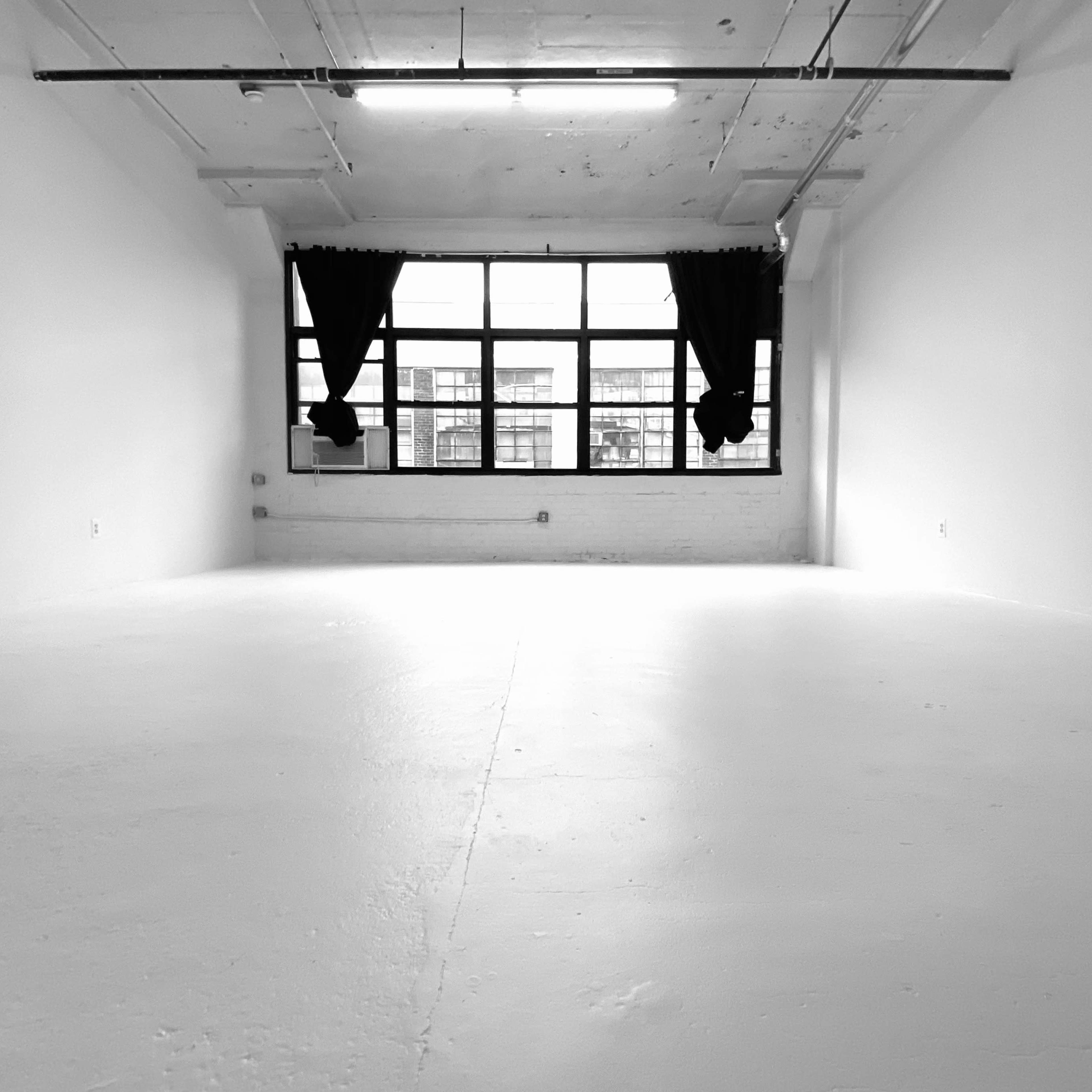 A large, empty room with white walls and a concrete floor features a row of windows covered by black curtains.