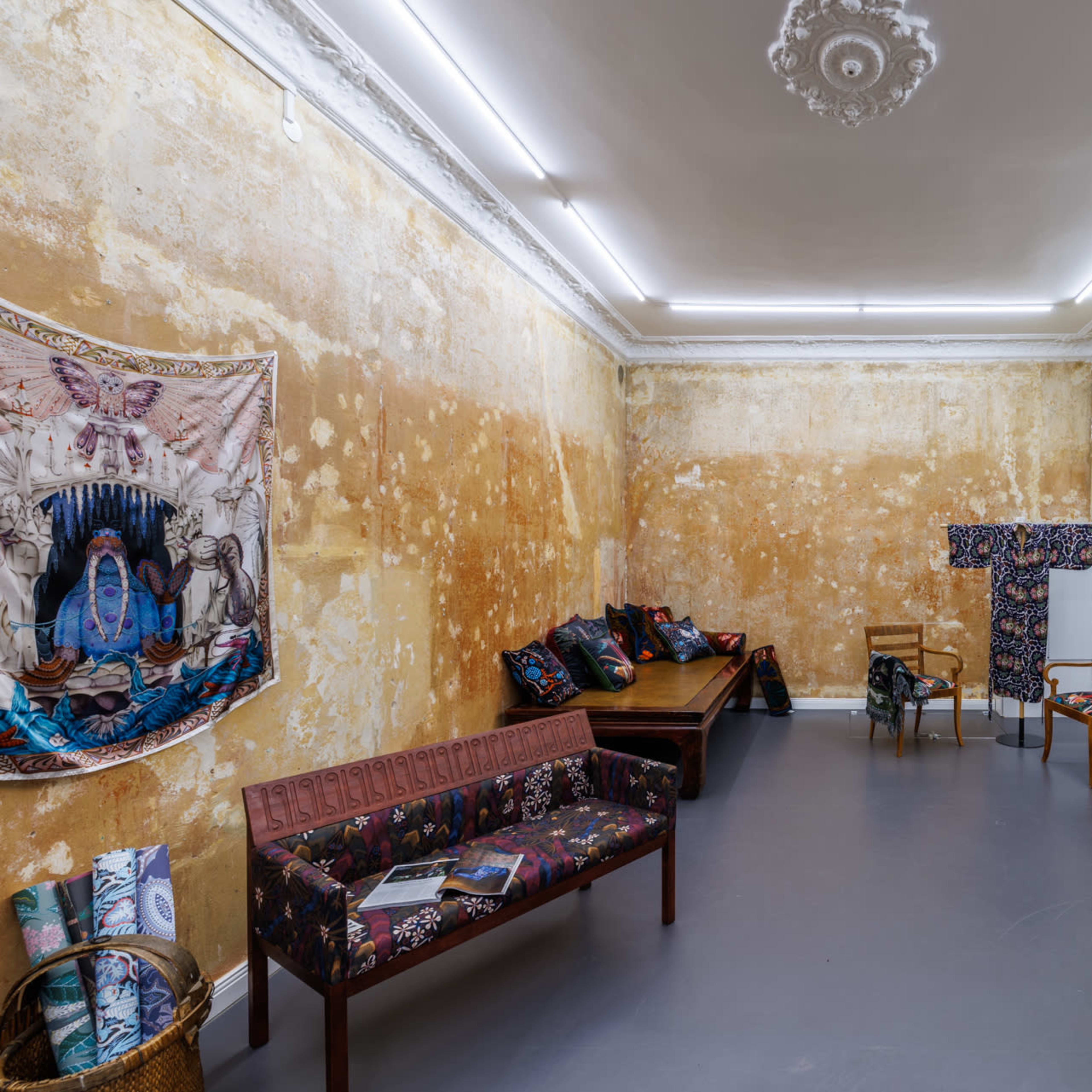A room with peeling yellow walls, featuring patterned fabric artworks, a wooden bench, chairs, and a decorative vase.
