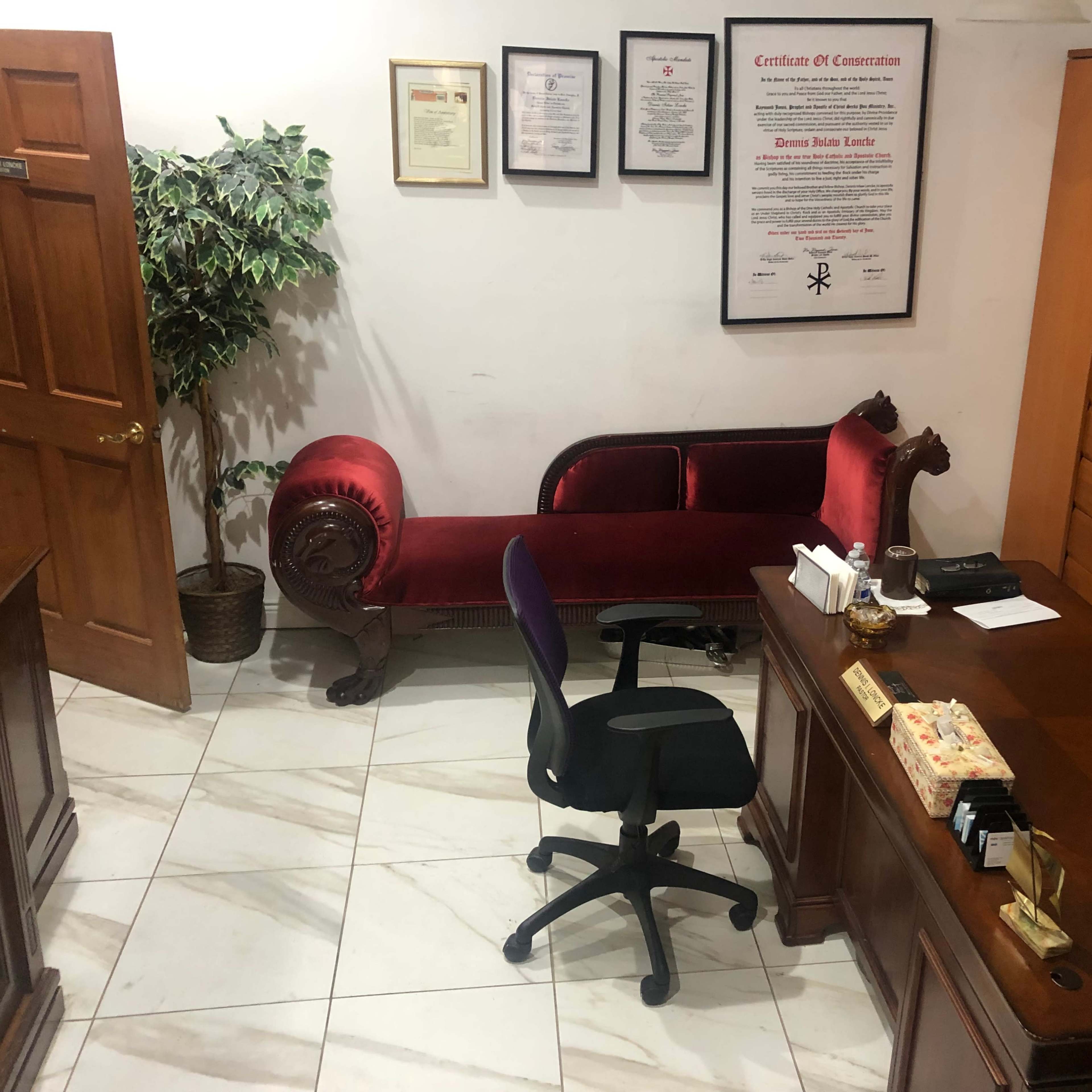 An office with a wooden desk, an office chair, a red upholstered sofa, and a decorative plant in the corner.