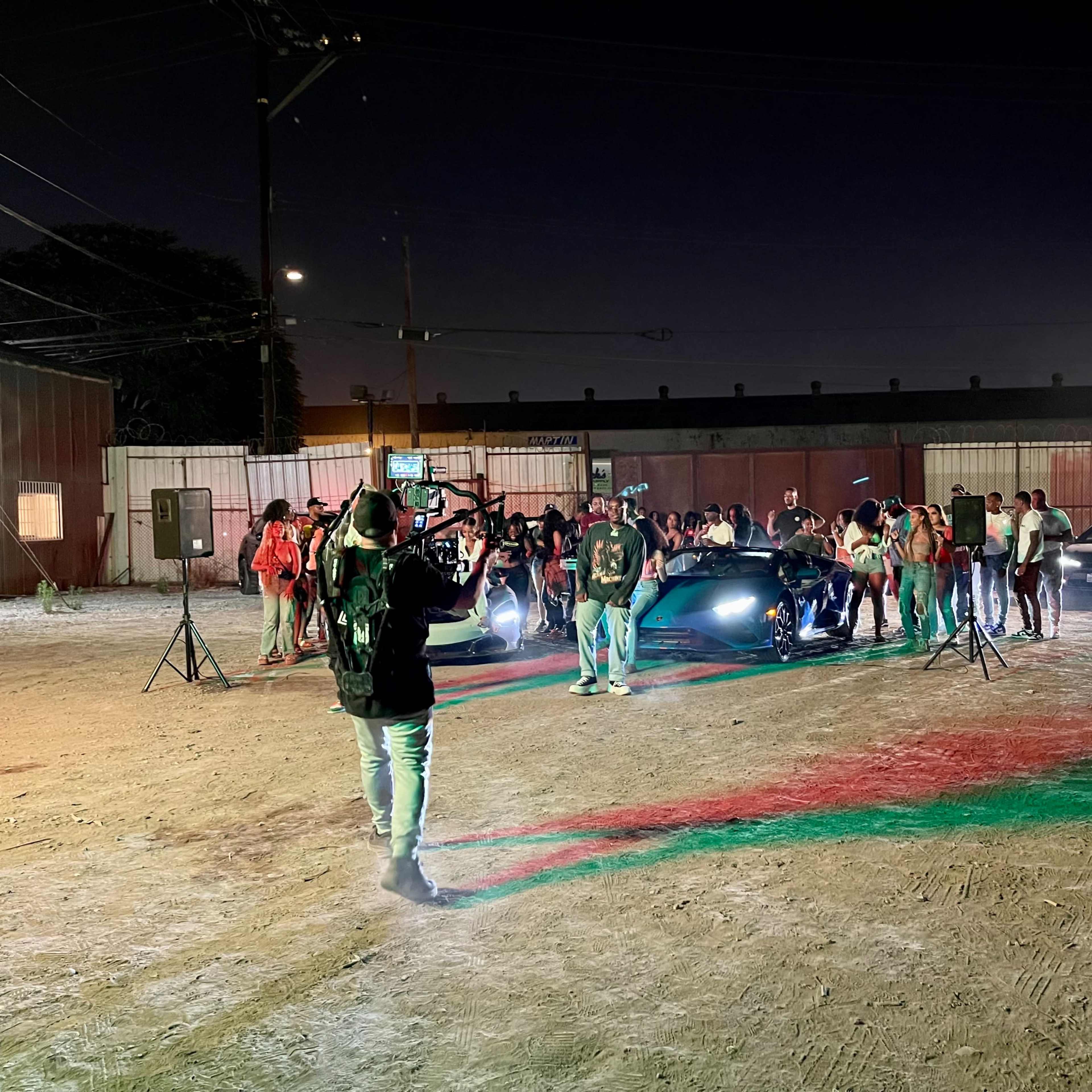 A group of people gathers around luxury cars in a lot illuminated by various lights at night.