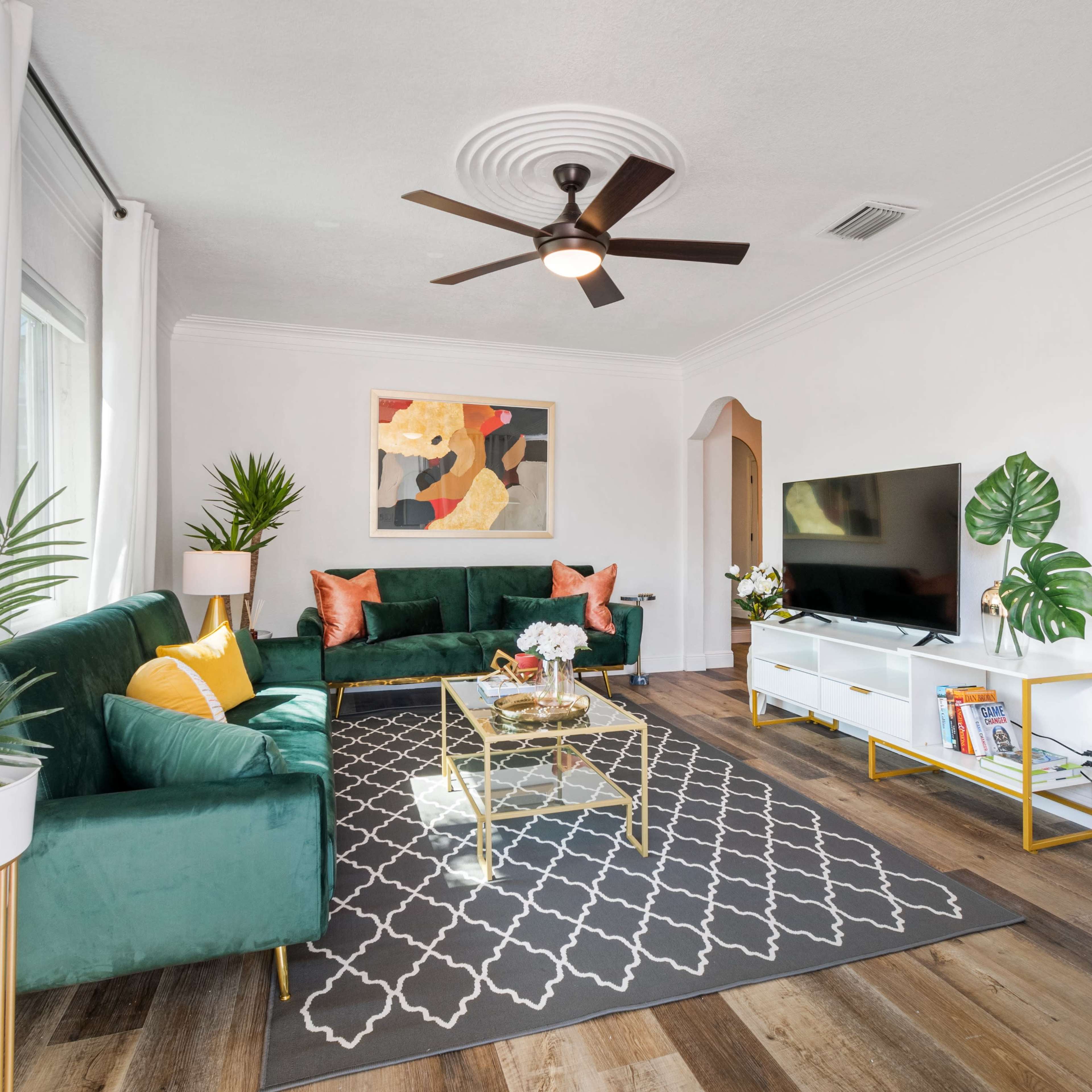 The living room features green sofas, a coffee table, a television cabinet, and decorative plants, arranged around a patterned area rug.