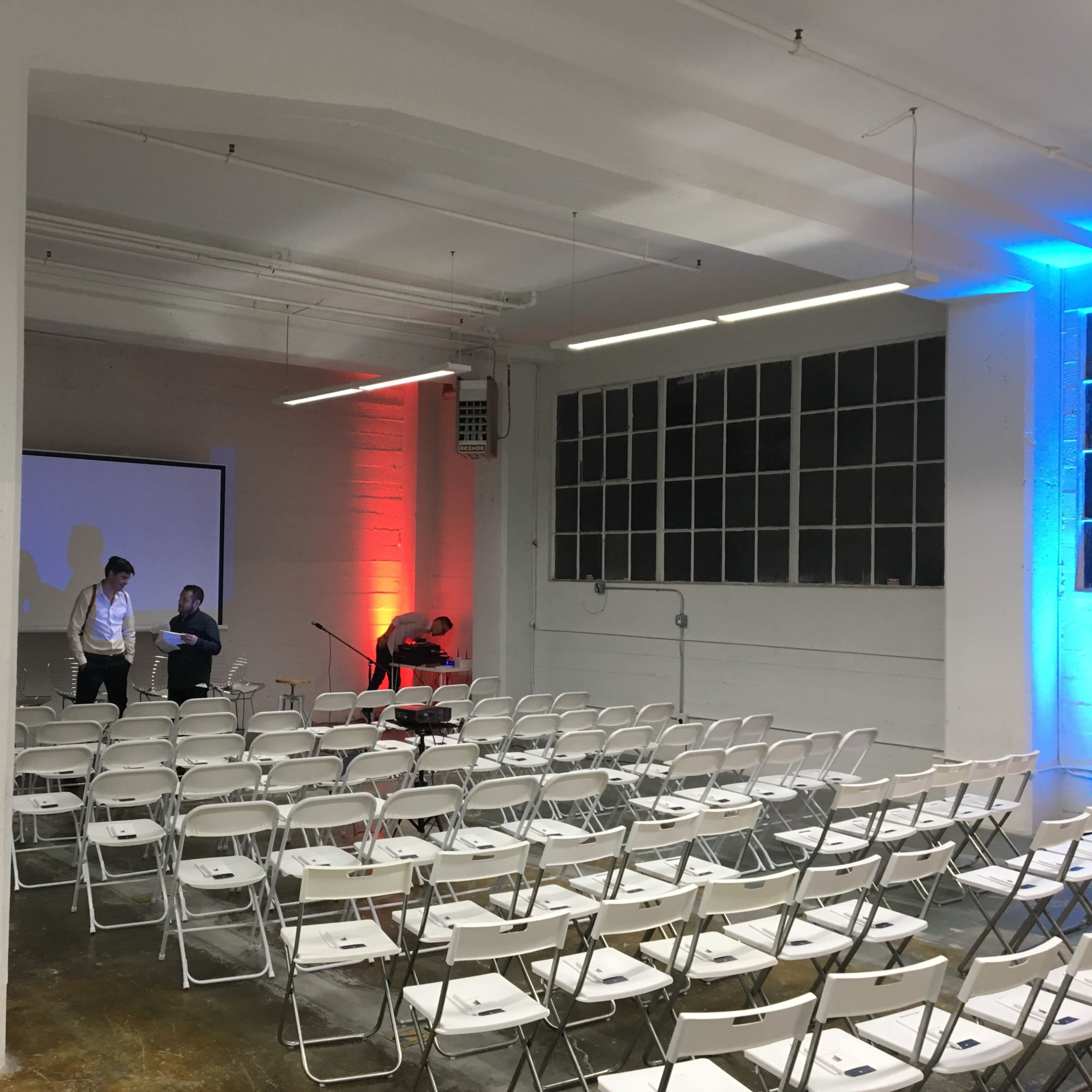 A large, empty room with white folding chairs arranged in rows facing a projector screen, where two people are preparing the setup.