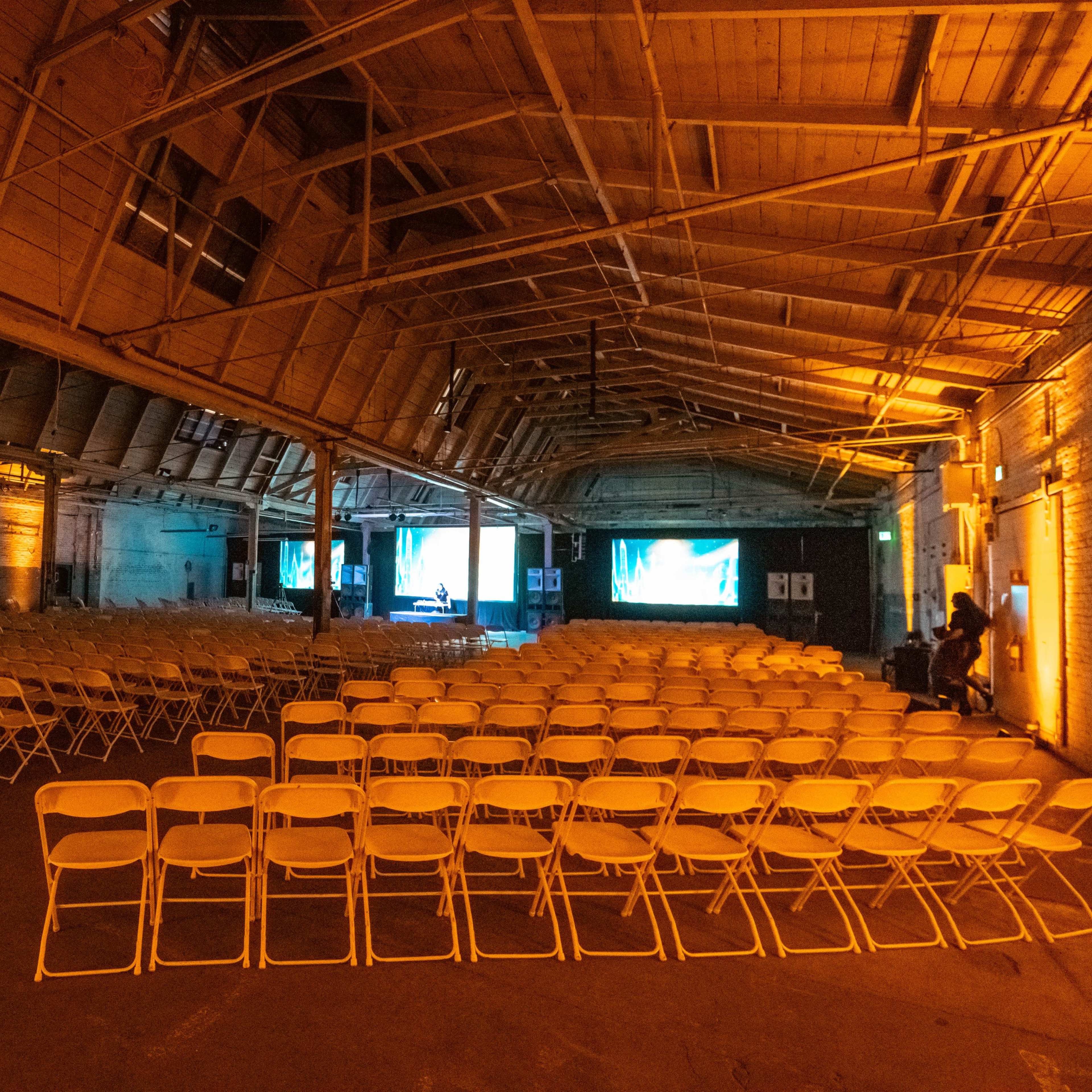 A spacious, empty venue with rows of white folding chairs set up in front of large screens.
