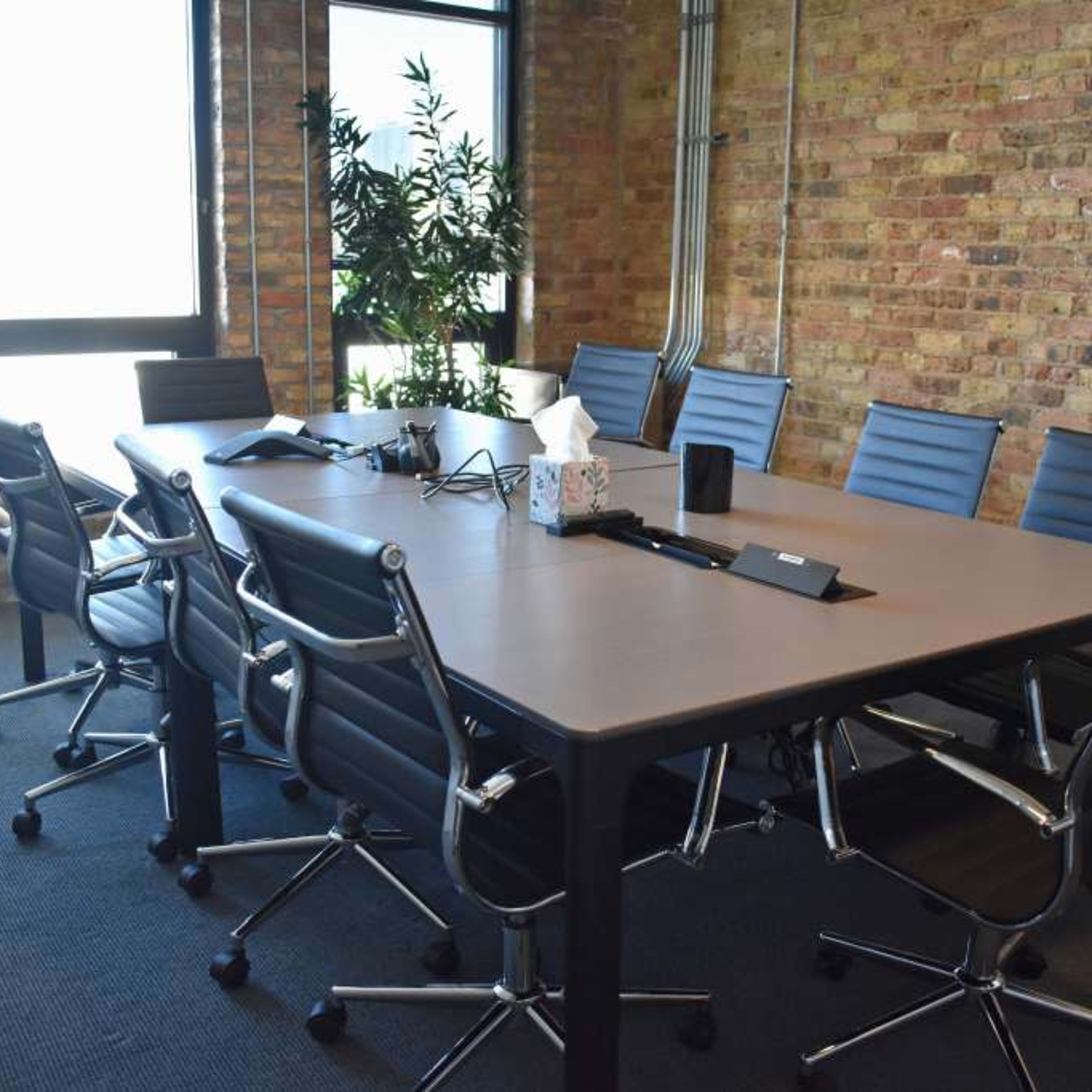 A modern conference room features a large rectangular table surrounded by ergonomic chairs, with windows and exposed brick walls.