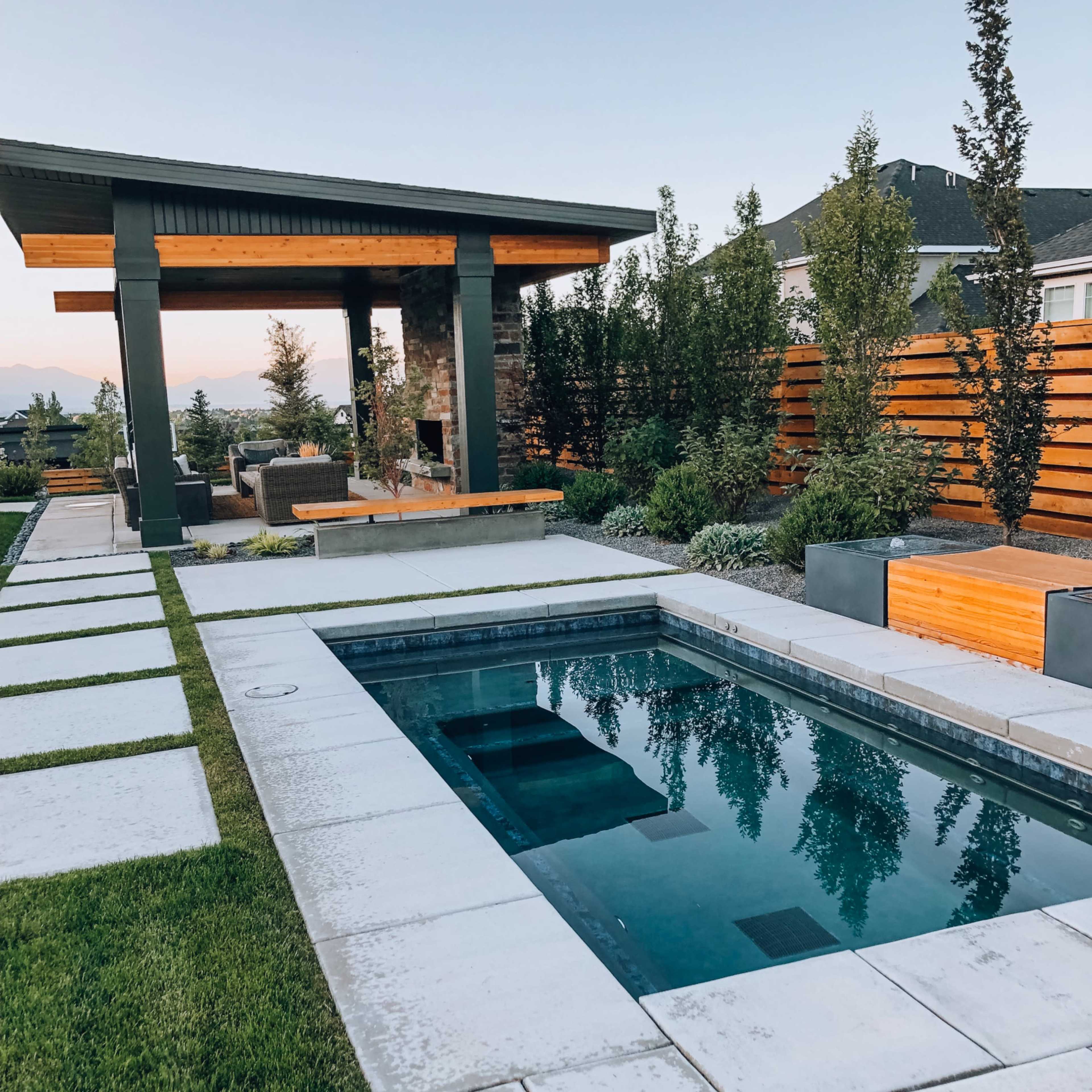A modern backyard featuring a rectangular swimming pool, a stone patio with seating, and landscaped greenery surrounding a pergola.