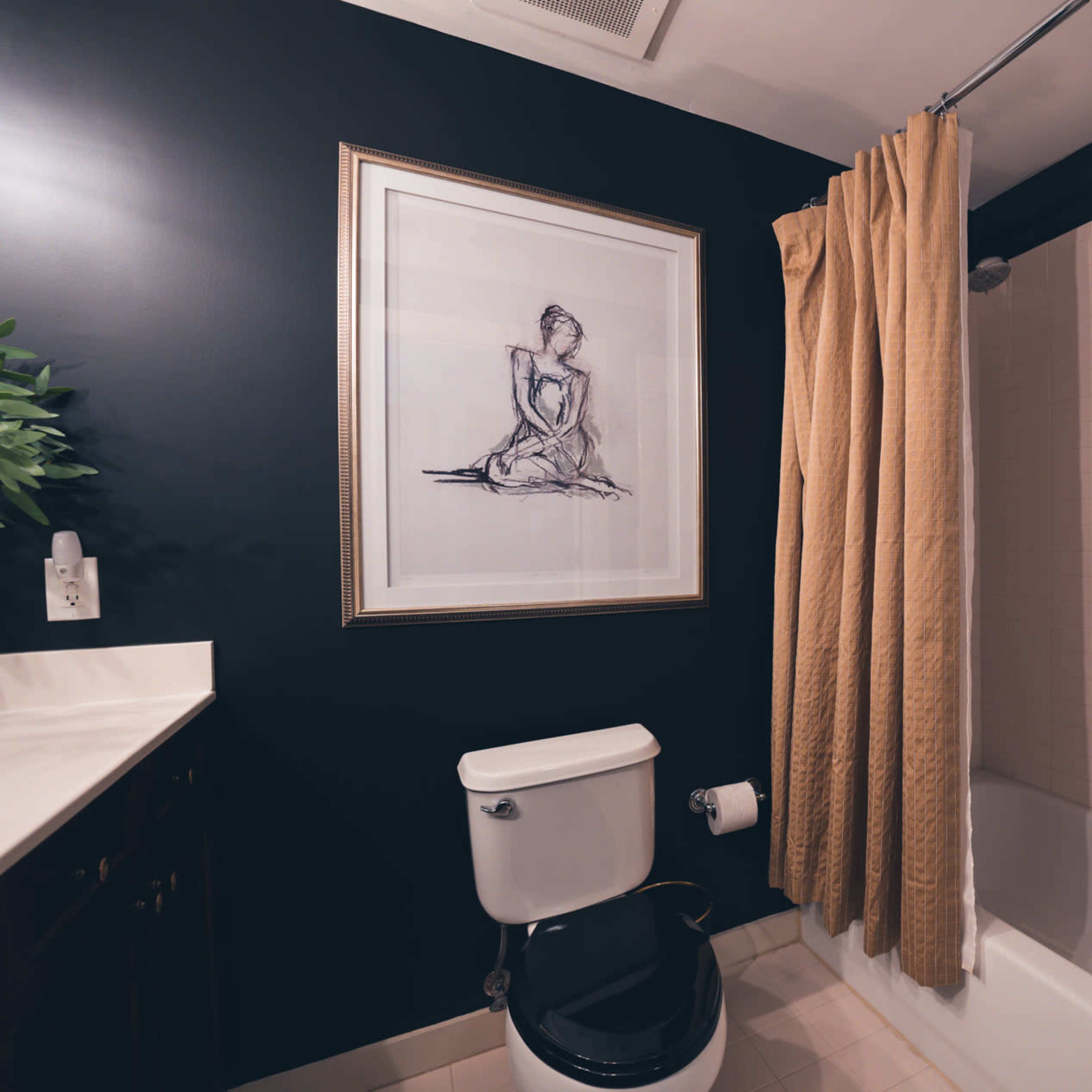 A bathroom with a dark wall, a shower with a curtain, a framed drawing, a toilet, and a light-colored floor.