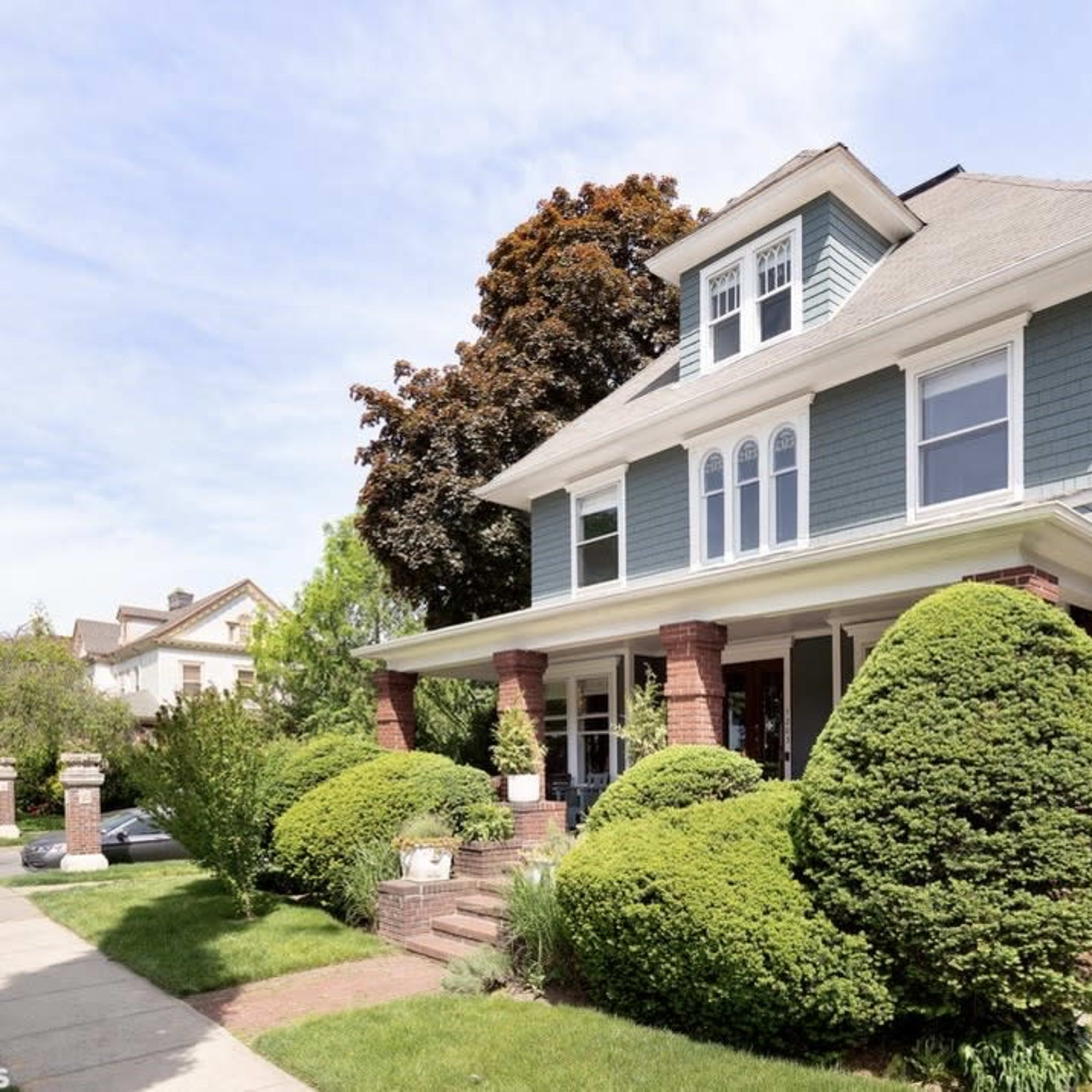 A three-story blue house with white trim, surrounded by well-maintained shrubs and trees.