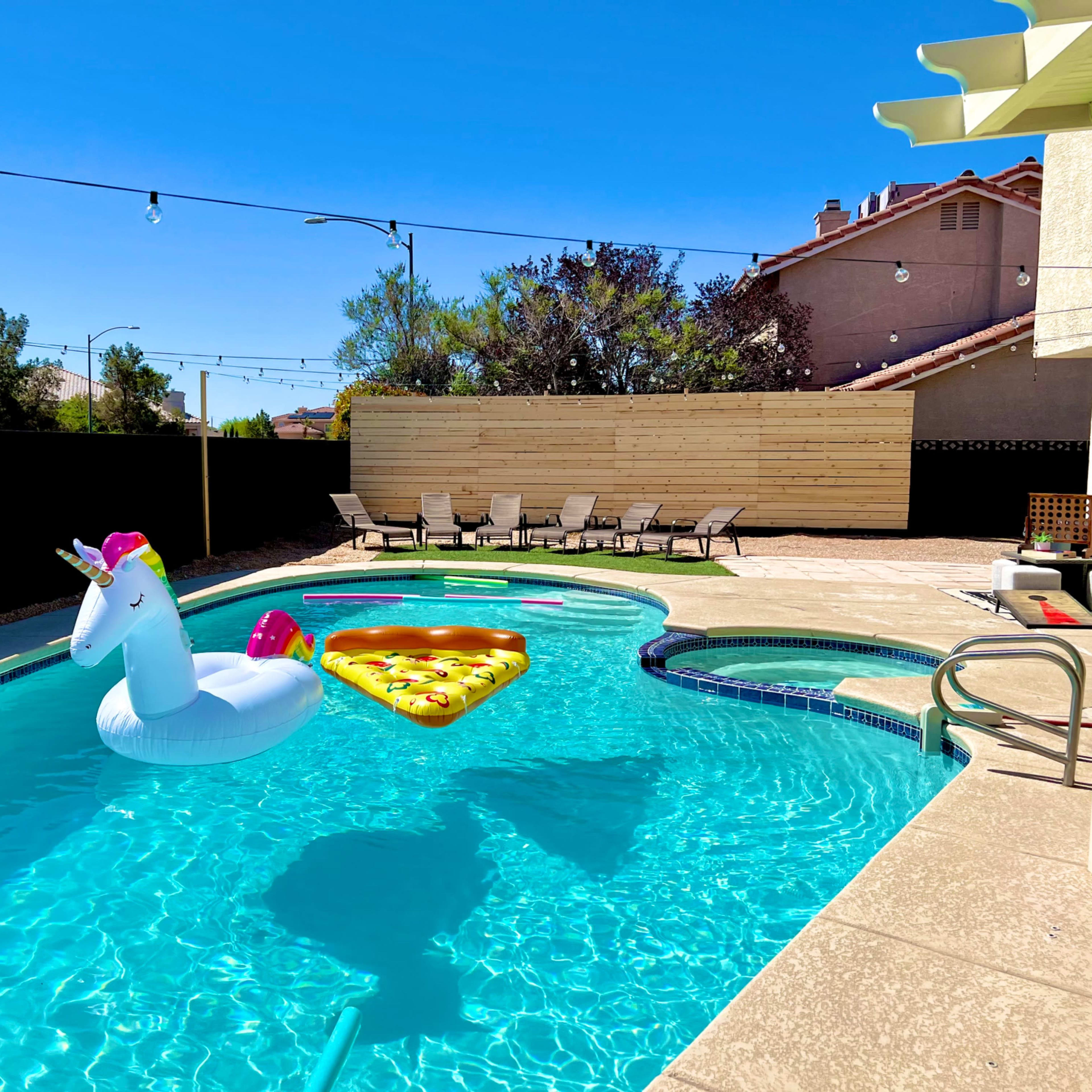 A unicorn-shaped inflatable and a pizza-shaped float are positioned in a swimming pool surrounded by lounge chairs and a wall.