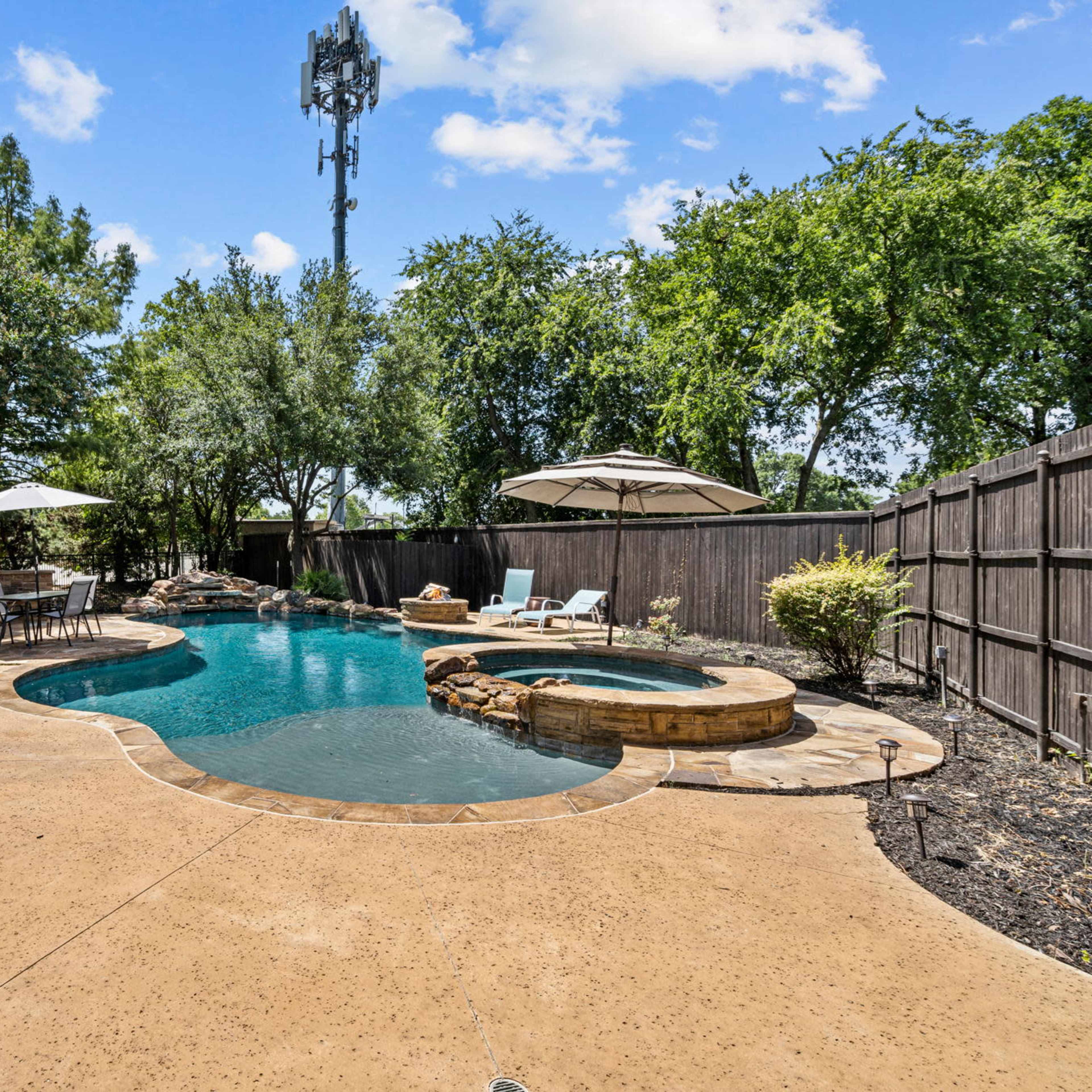 A backyard pool area featuring a swimming pool, a hot tub, lounge chairs, and surrounding greenery.