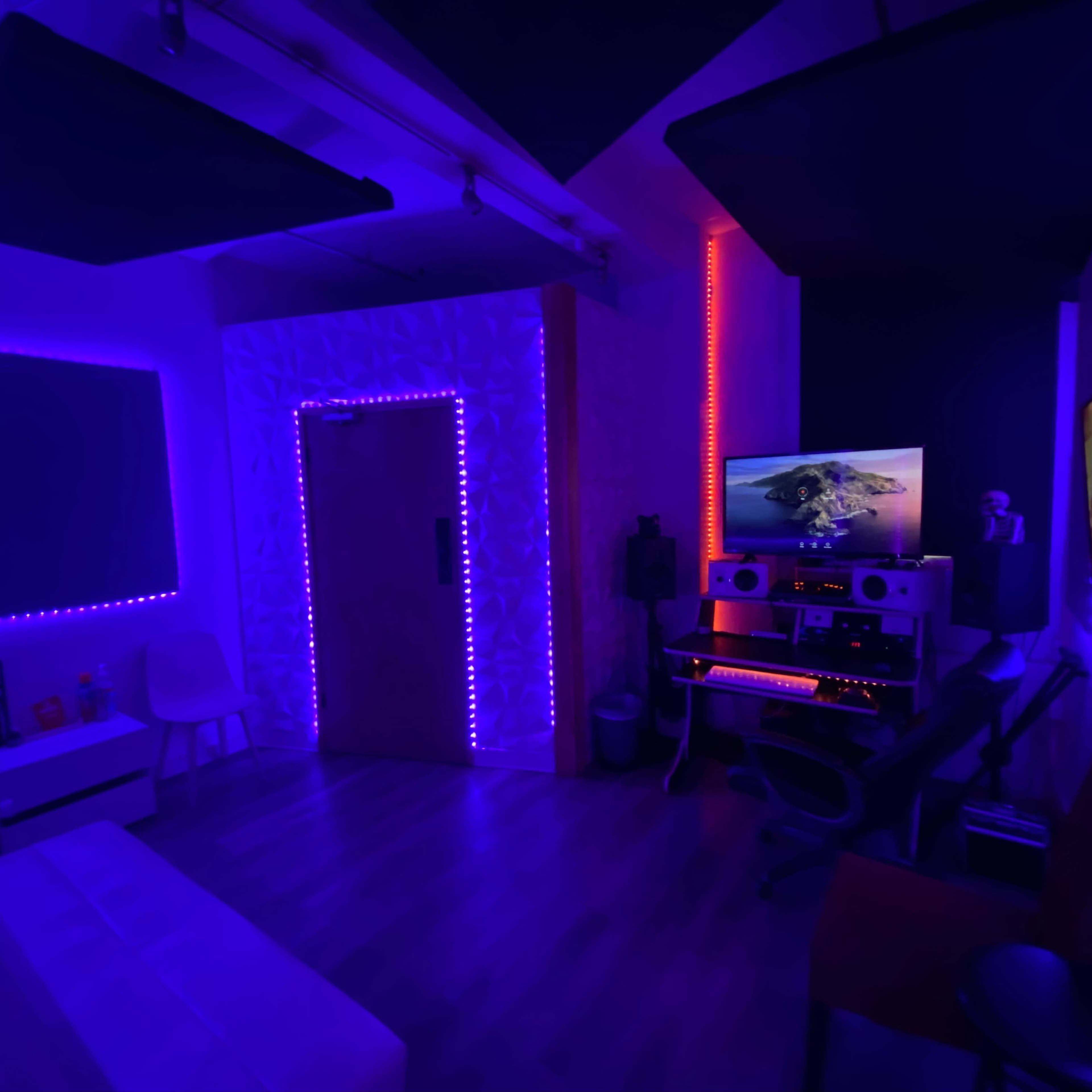 A dimly lit room with purple and orange LED lights, featuring a desk with audio equipment, a computer monitor, and a seating area.