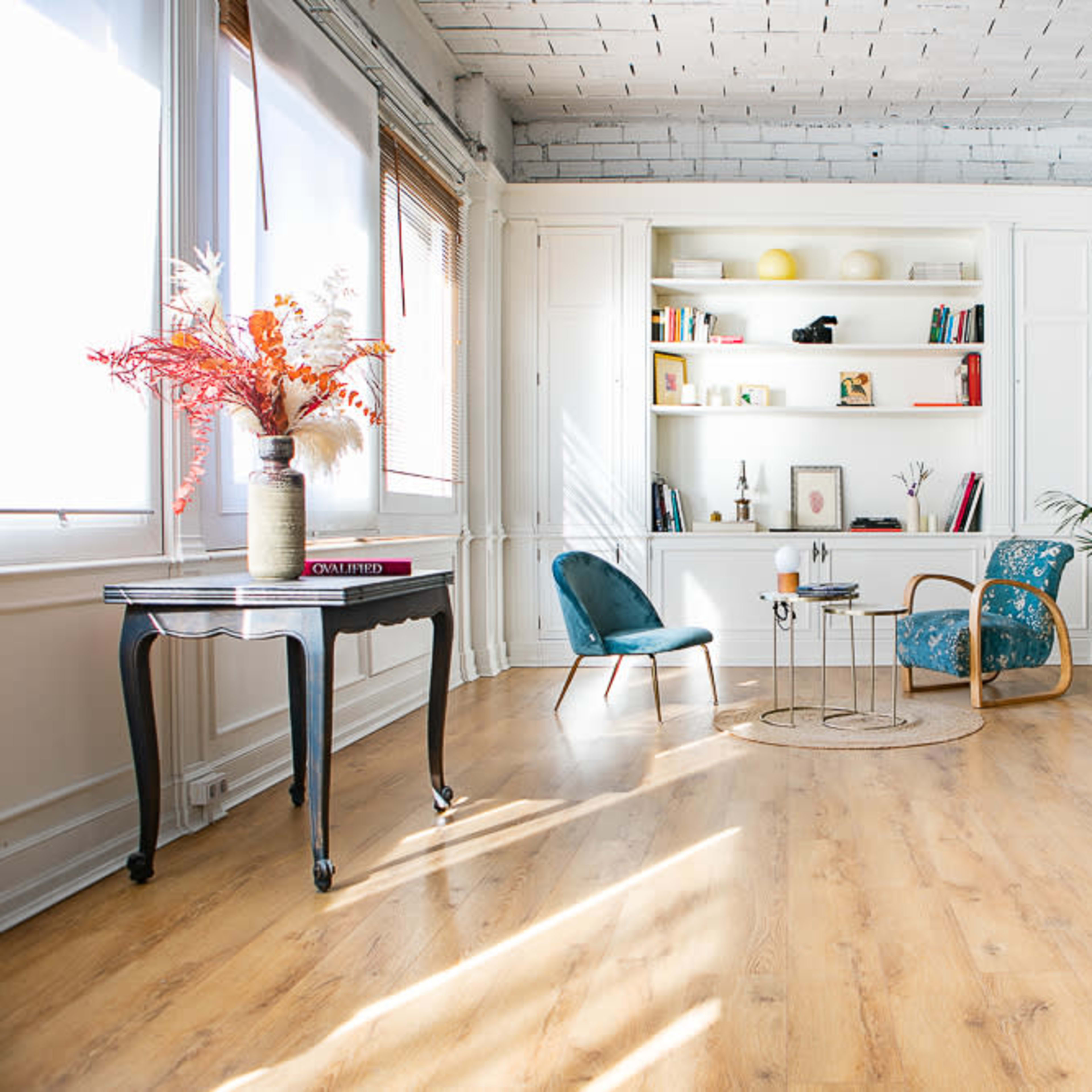A bright, modern living space with a light wooden floor, a bookshelf filled with books and decorative items, and several stylish chairs arranged around a small table.