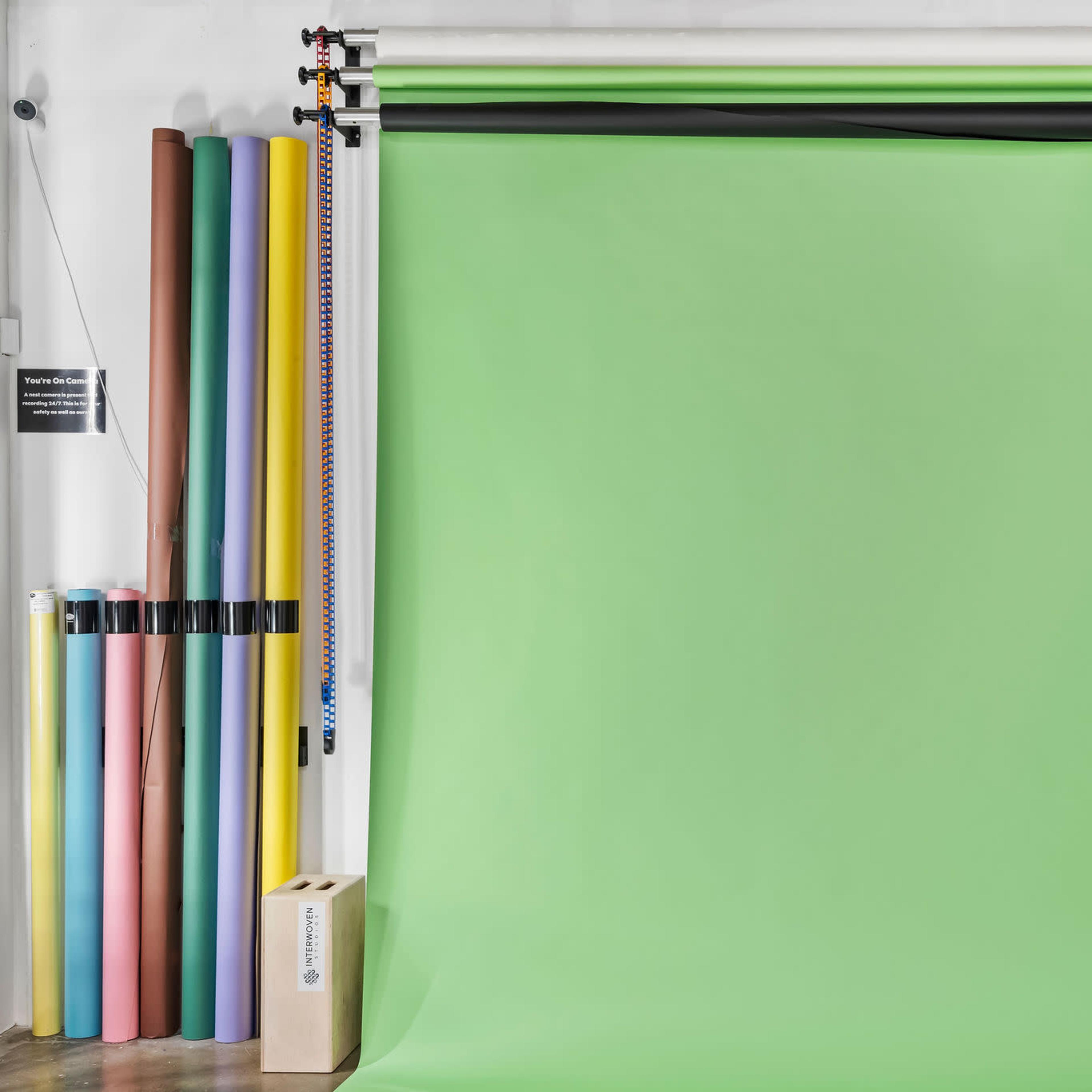 A photography studio with a green backdrop, various colored paper rolls on the left, and a small plant in a corner.