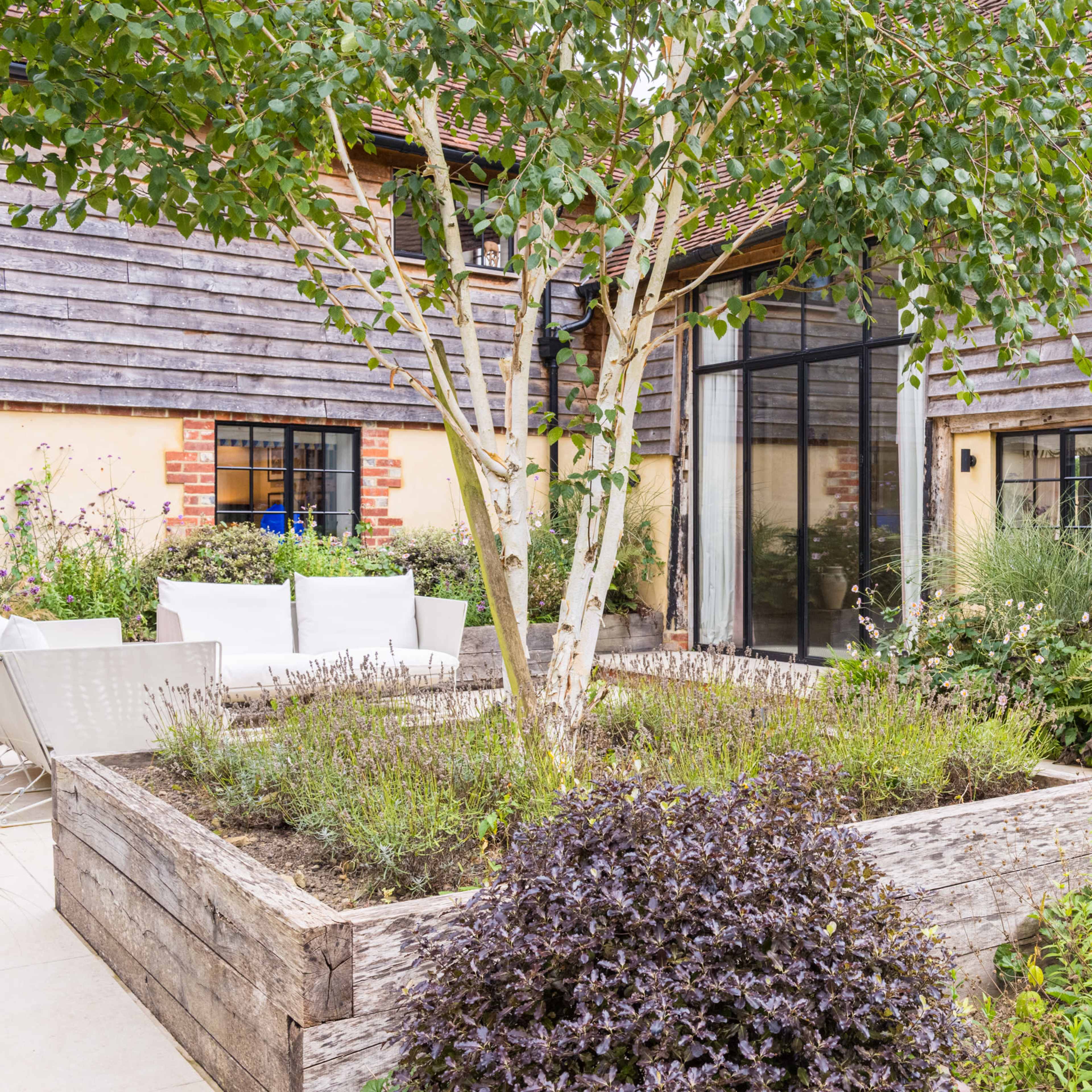 A neatly landscaped garden features a wooden planter box with various plants, surrounded by a rustic-style building with sliding glass doors and a trees in the center.