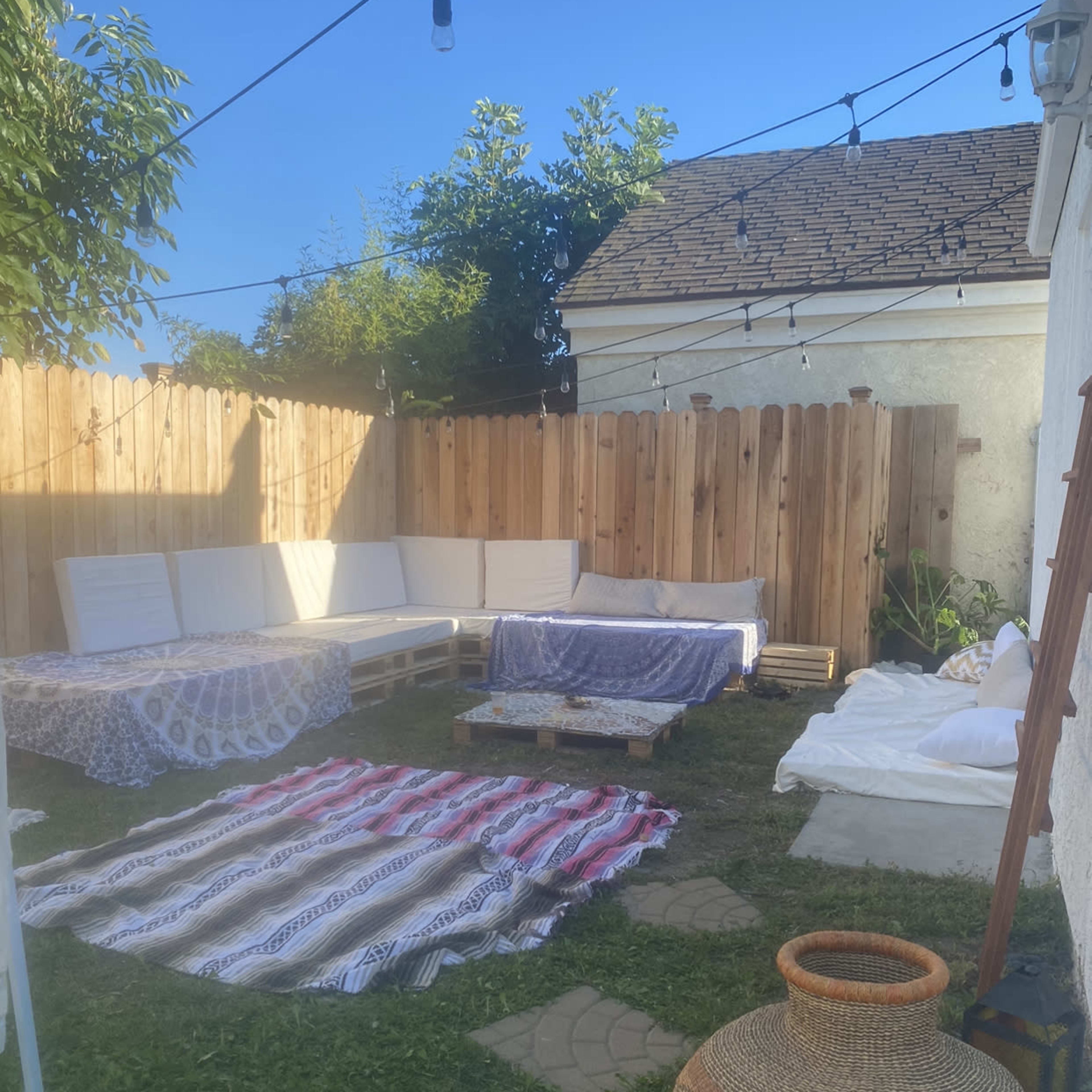 A backyard space features pallet seating covered with fabrics and scattered blankets on the grass, bordered by a wooden fence and strung with lights overhead.