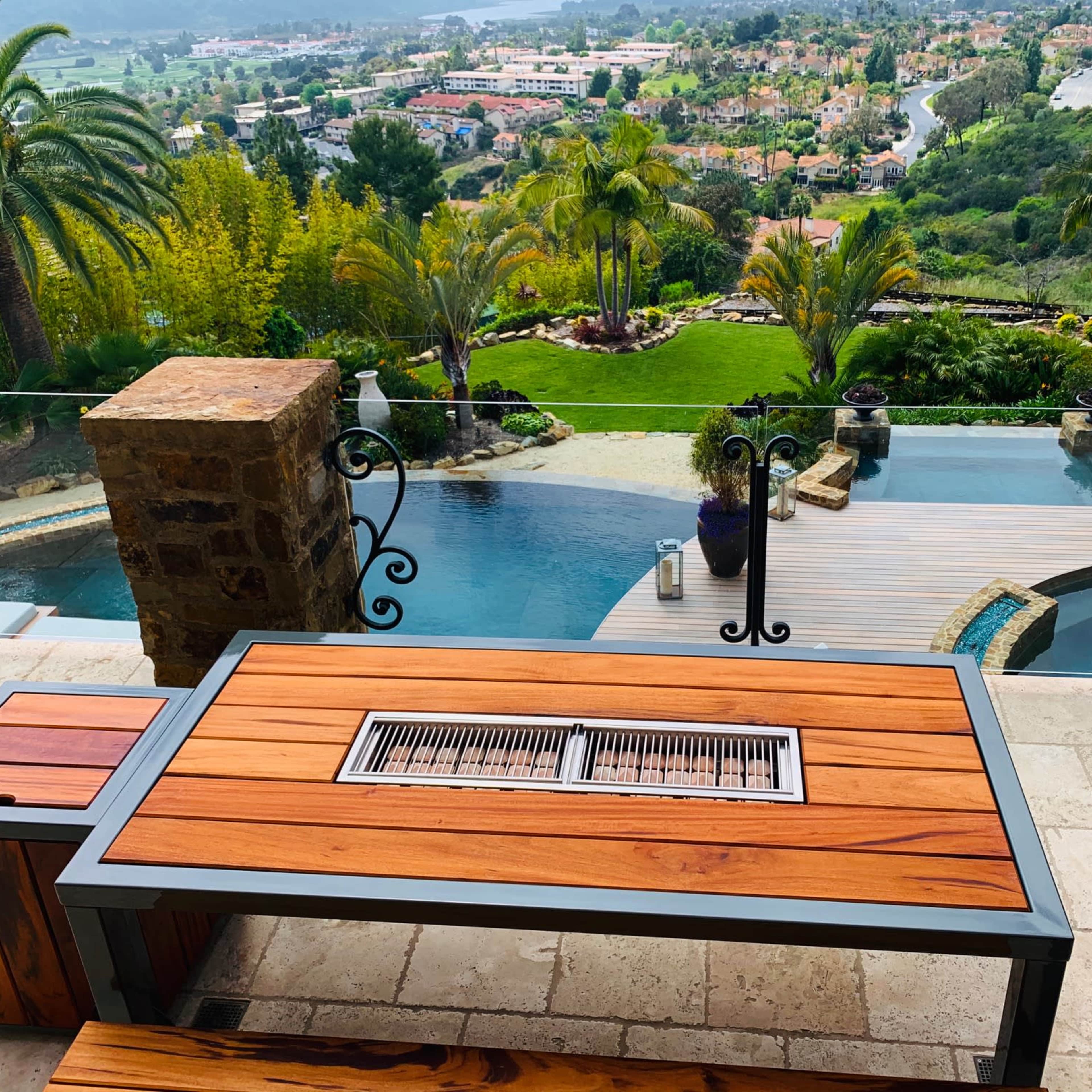 A wooden table with a grill sits on a stone patio overlooking a pool and a landscaped view of hills and homes.