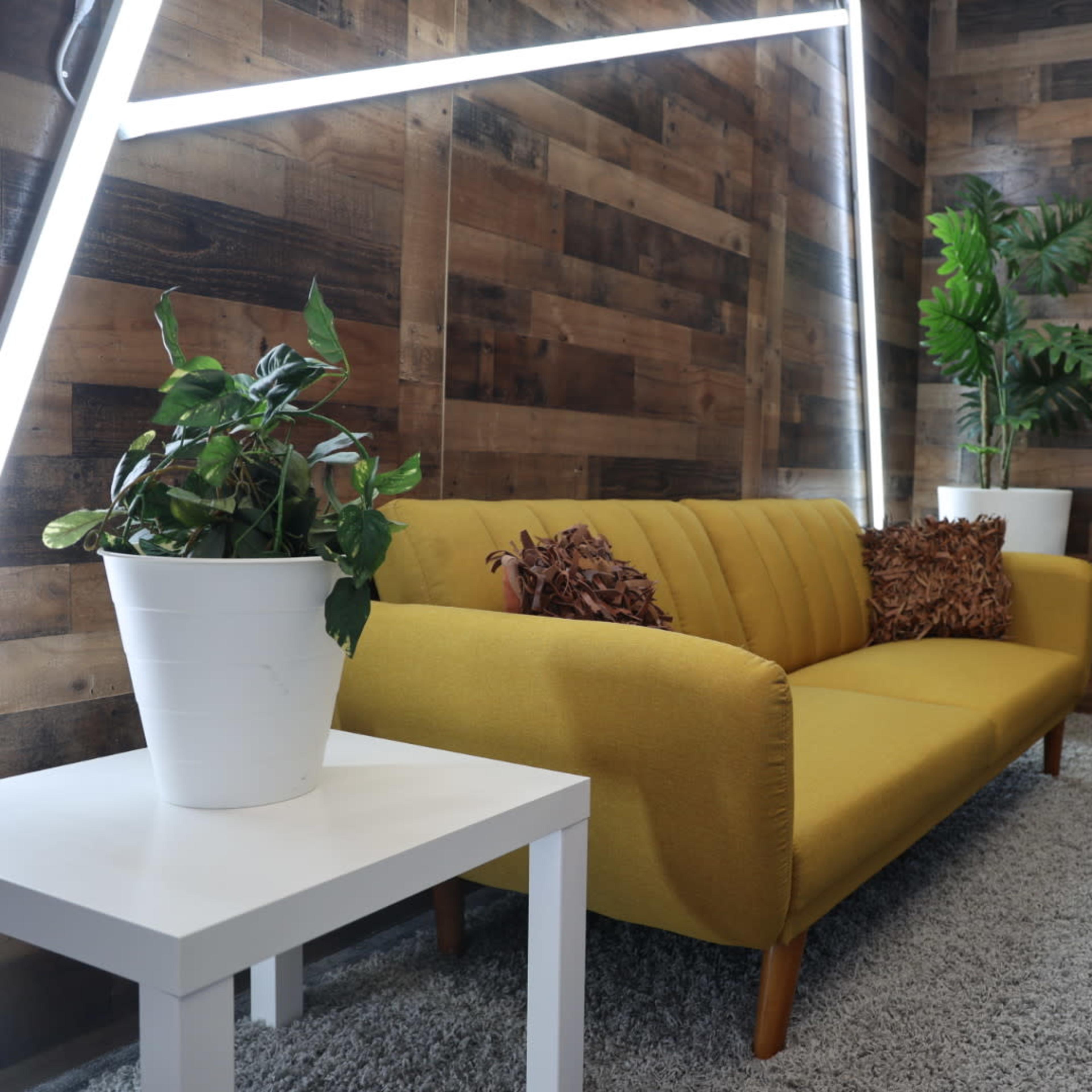 A yellow couch with decorative pillows sits next to a white table and potted plants in a room with wooden walls and gray carpet.