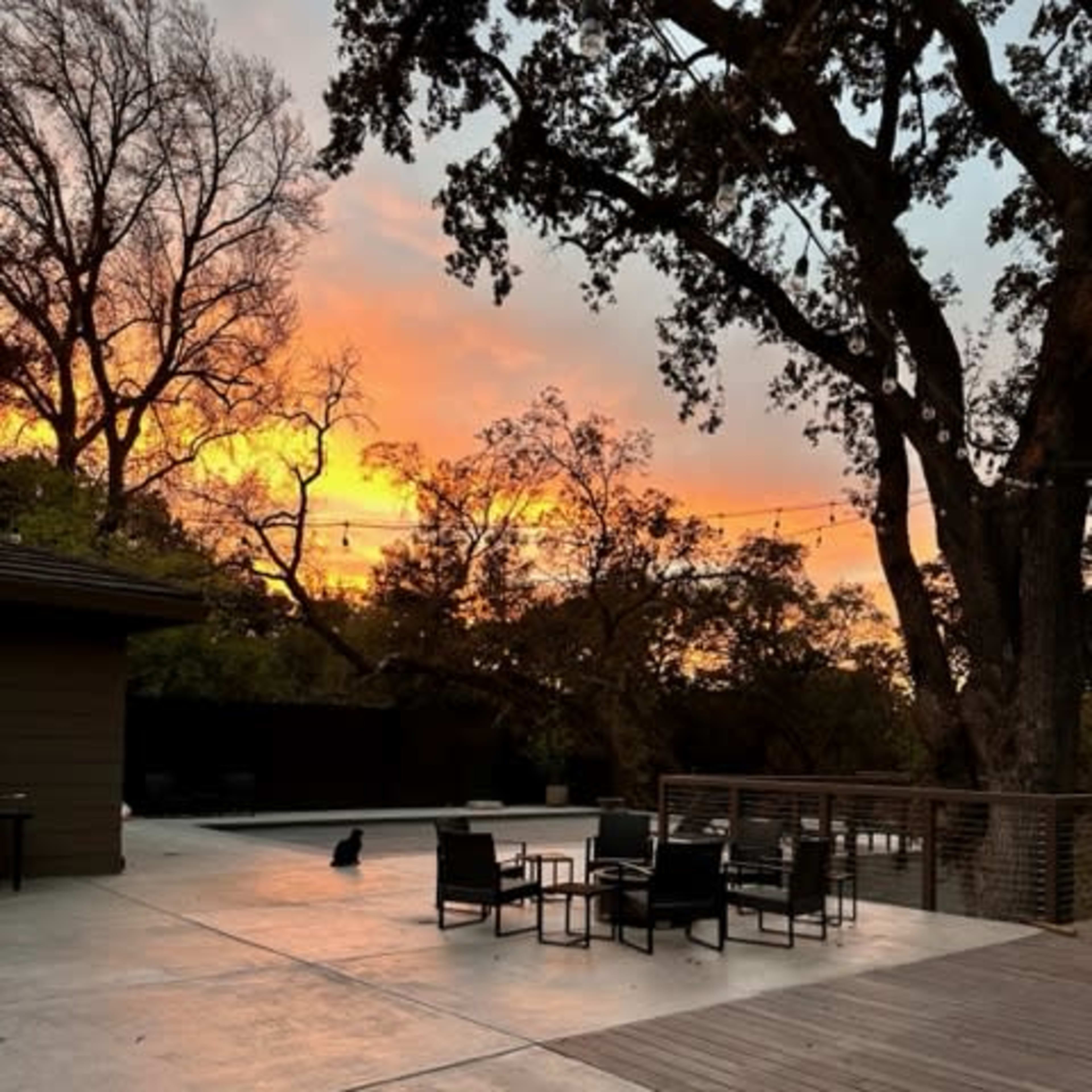 A patio with seating arrangements sits under a vibrant sunset, surrounded by trees.