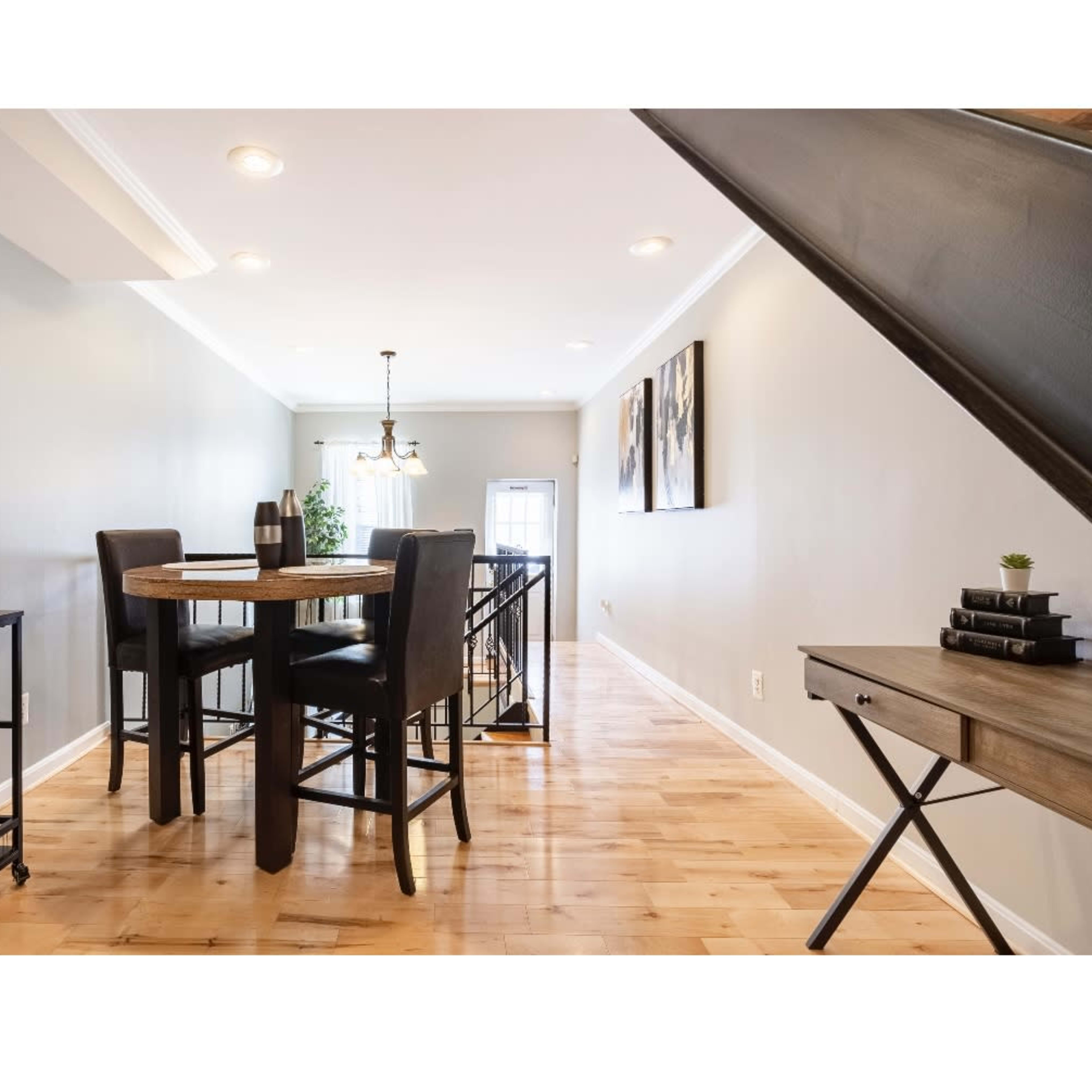 A dining area with a round wooden table and black chairs is positioned alongside a hallway featuring light wood flooring and gray walls.