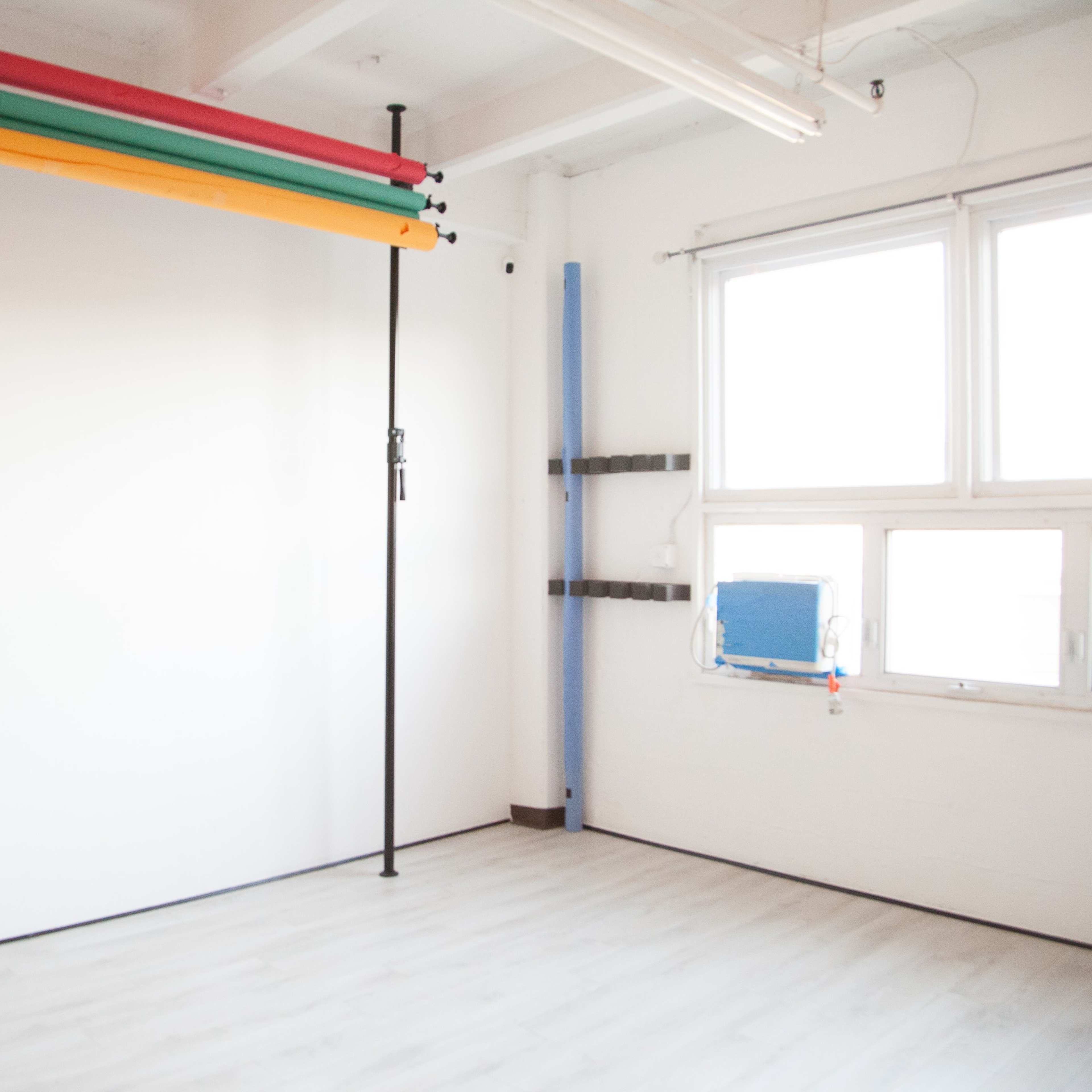 A bright, empty studio space with a blank white wall and colorful backdrops hanging on a vertical support.
