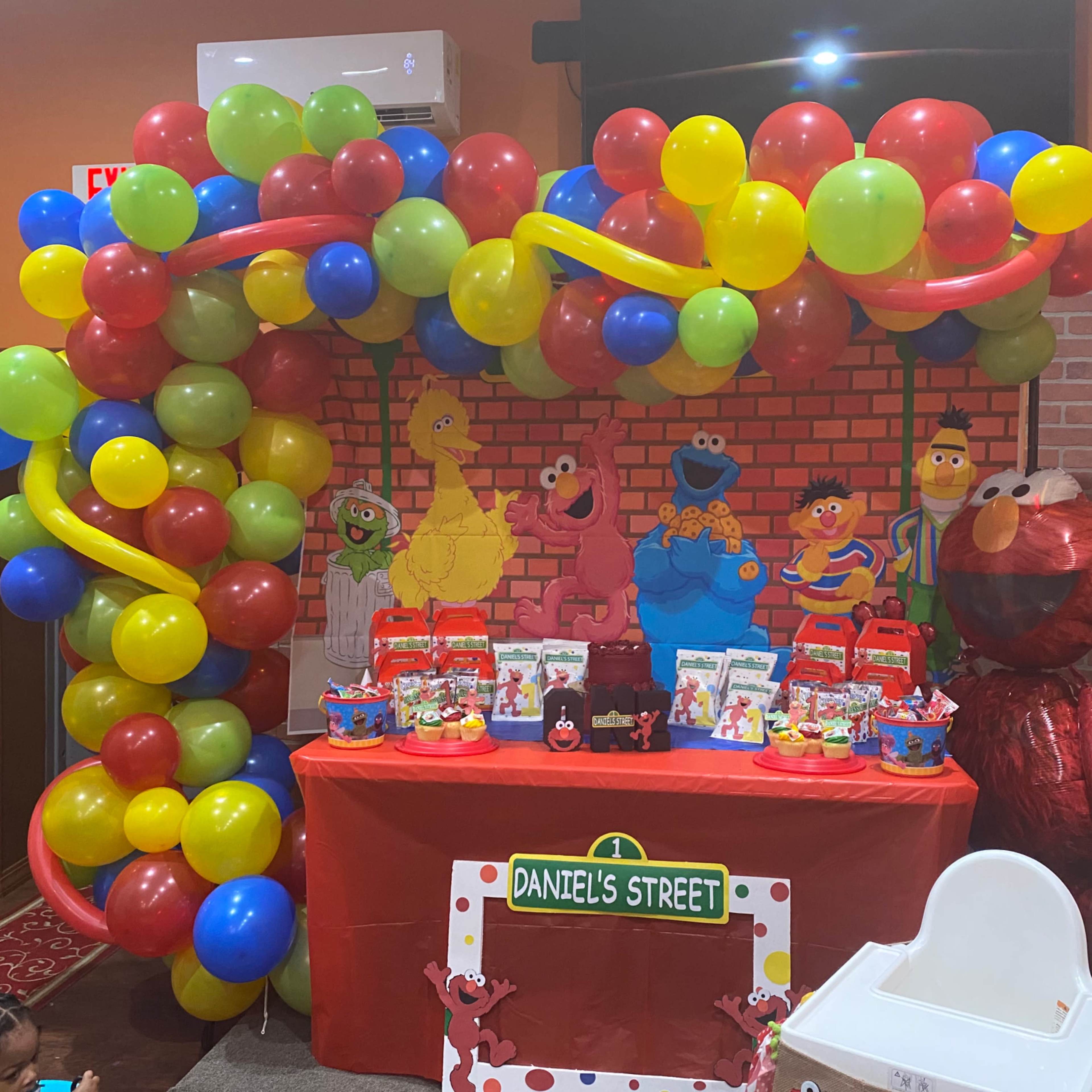 A colorful birthday party setup featuring a balloon arch, a table decorated with party favors, and a backdrop with Sesame Street characters.