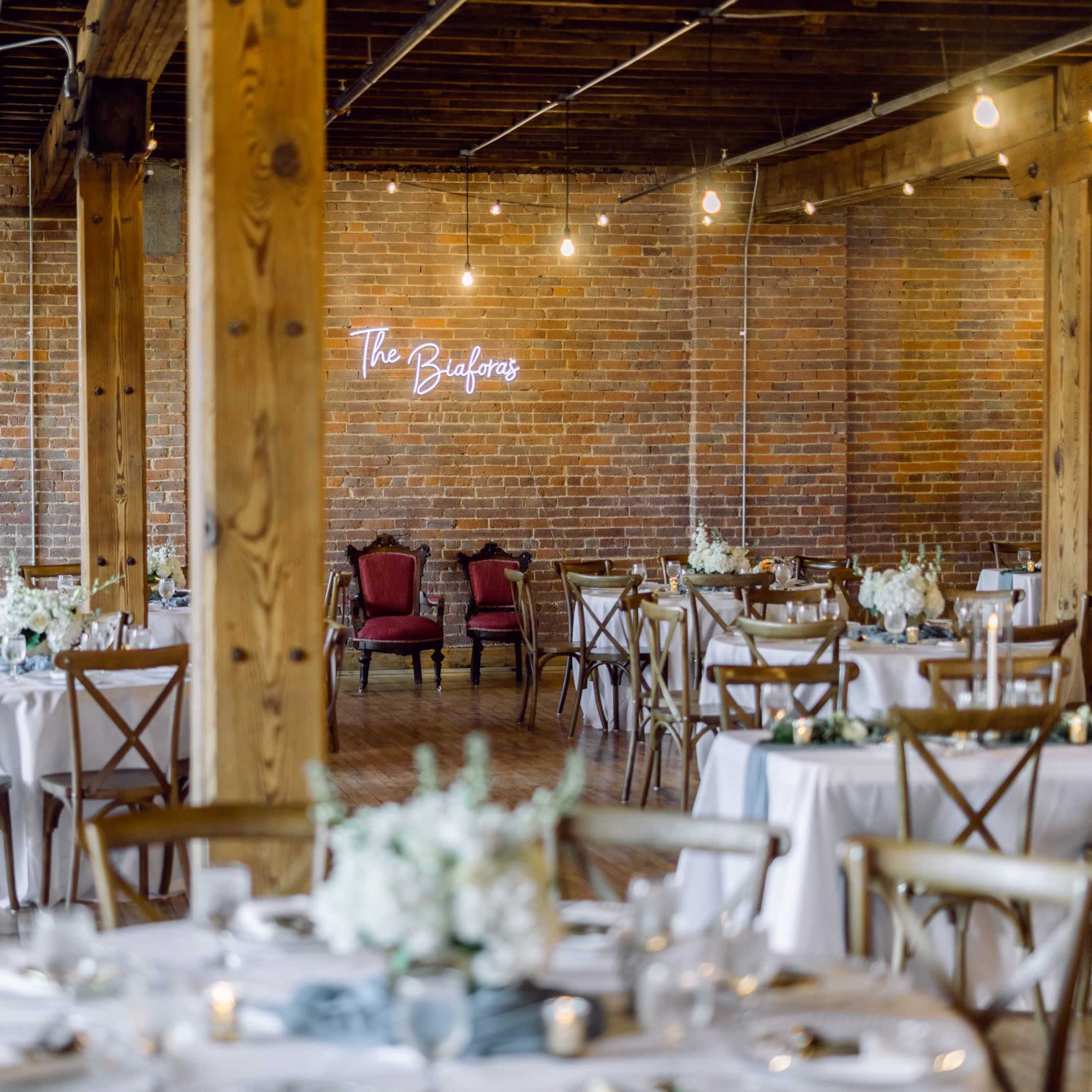 A spacious event venue with wooden beams, round tables set with white tablecloths, and a neon sign that reads "The Building."