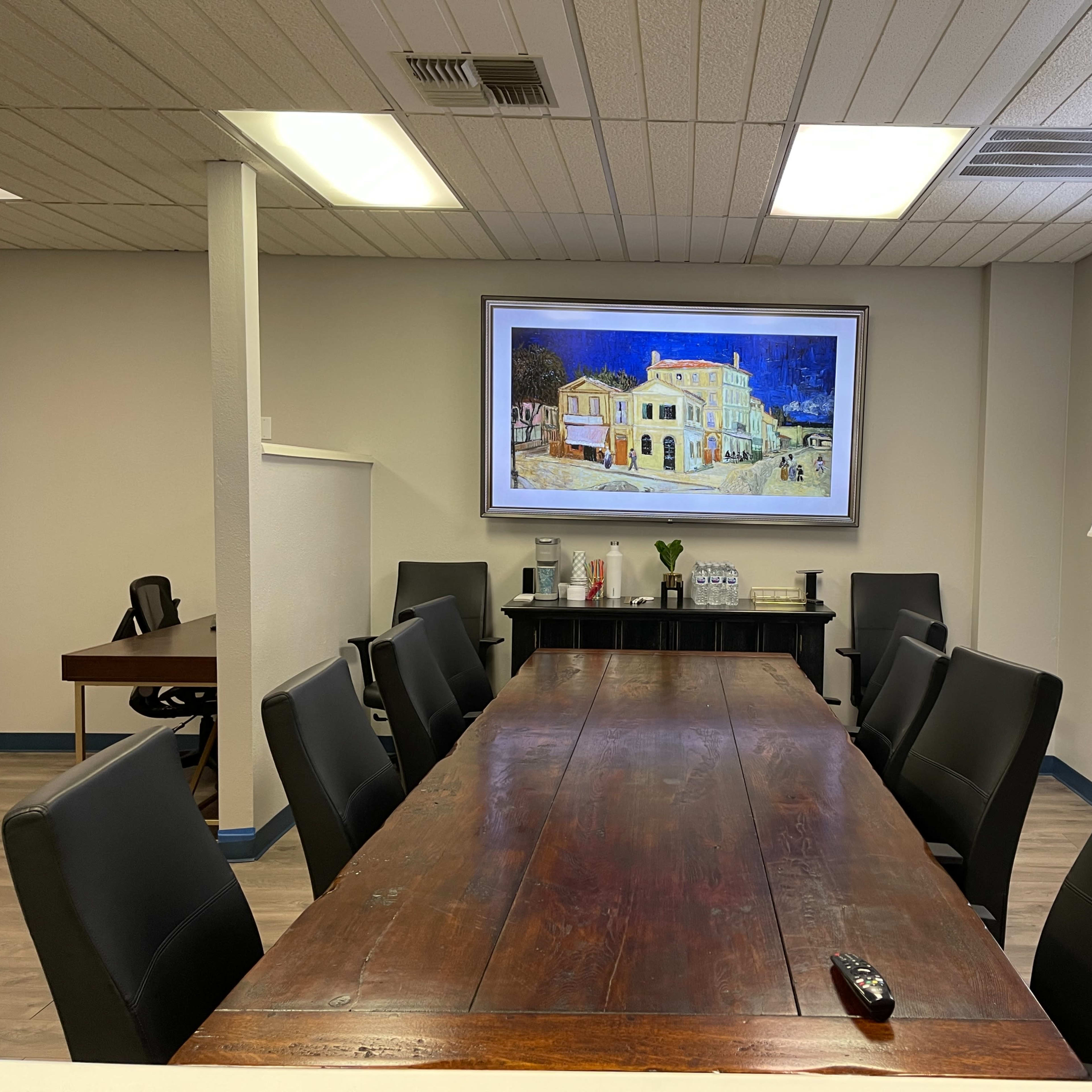 A long wooden conference table is set in a meeting room with black chairs, framed by a large painting and two lamps on opposite sides.