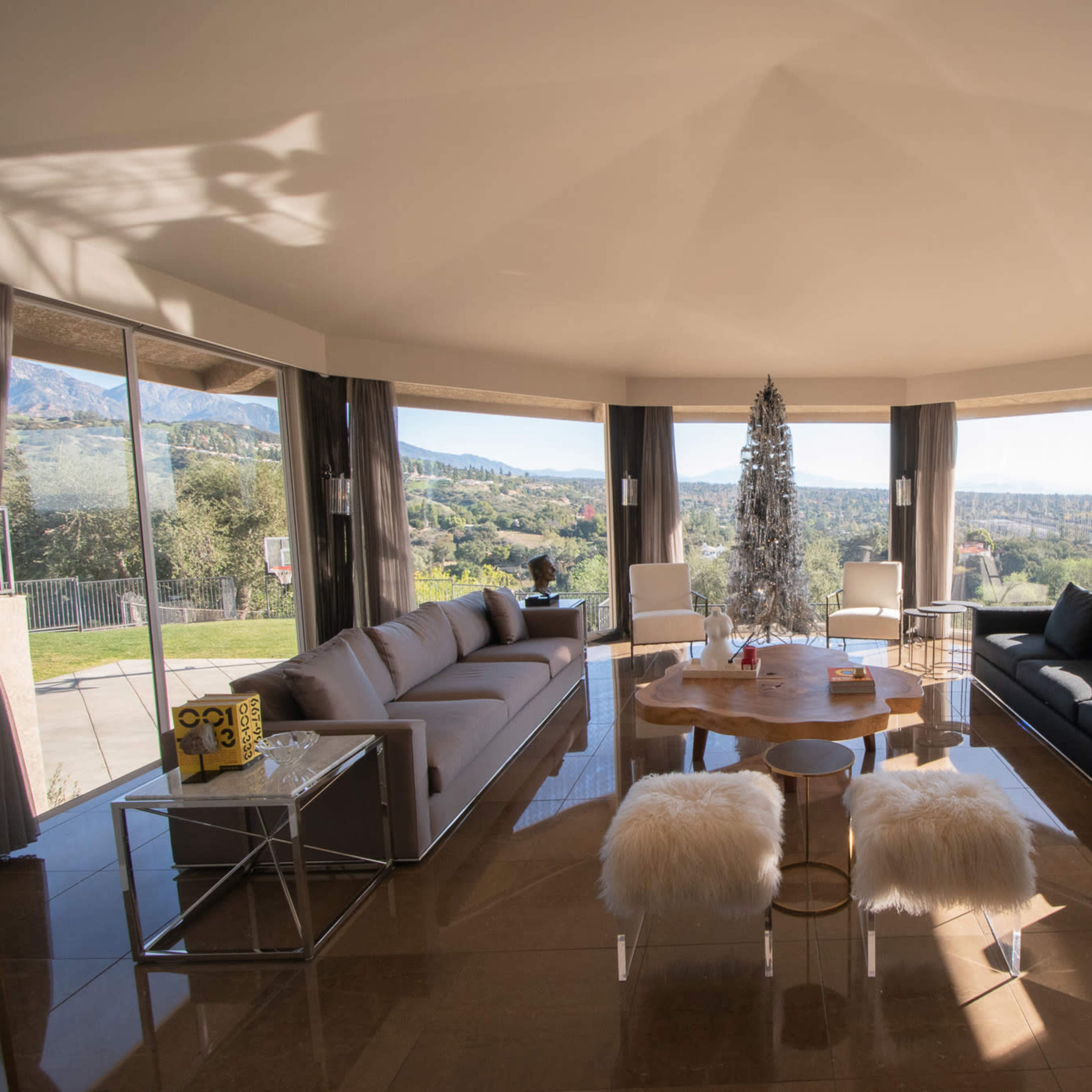 A modern living room with large windows offering a scenic view, featuring a round table surrounded by various seating arrangements and a decorative tree.