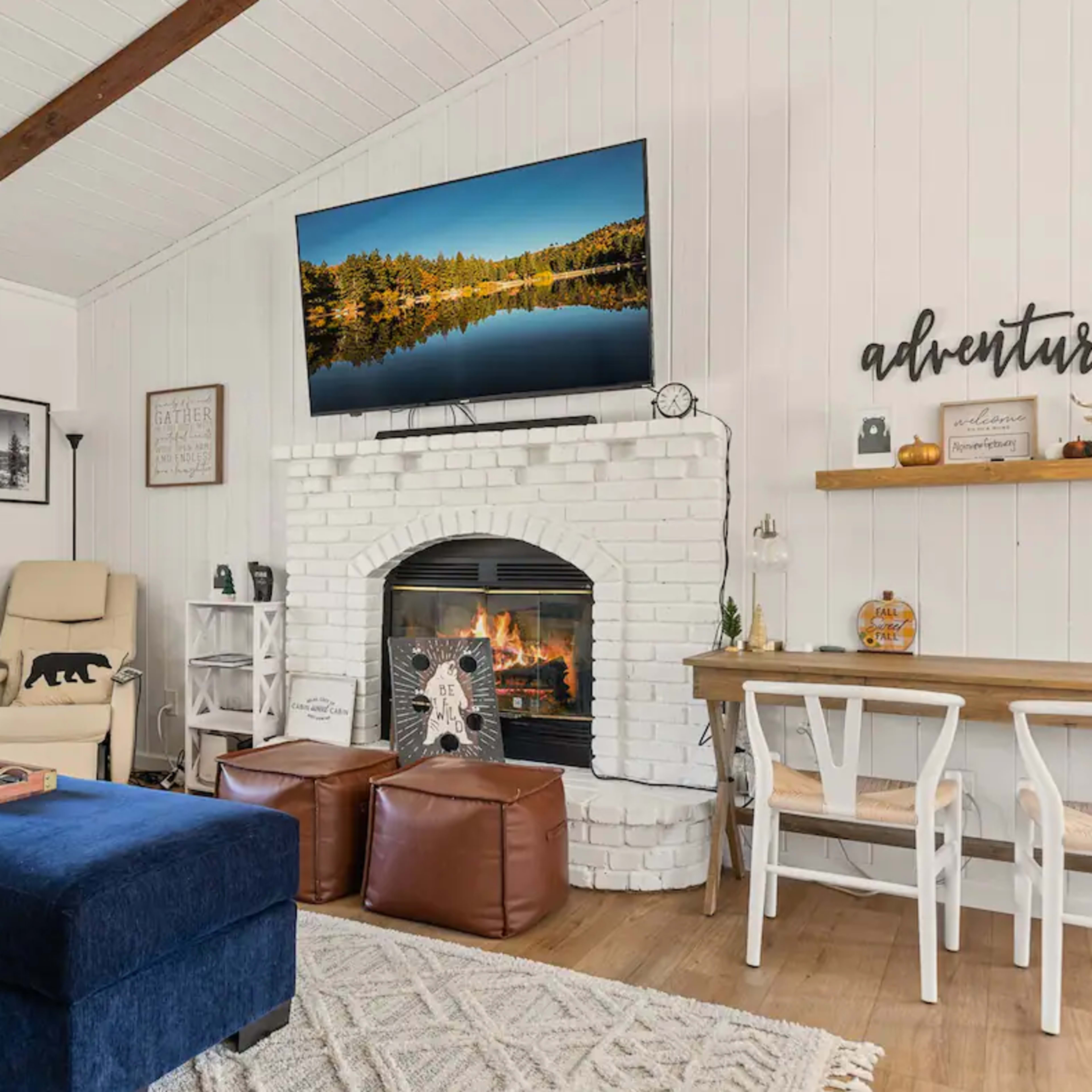 A cozy living room features a blue ottoman, a white brick fireplace, and a large television mounted above it, with comfortable seating and decorative elements throughout the space.