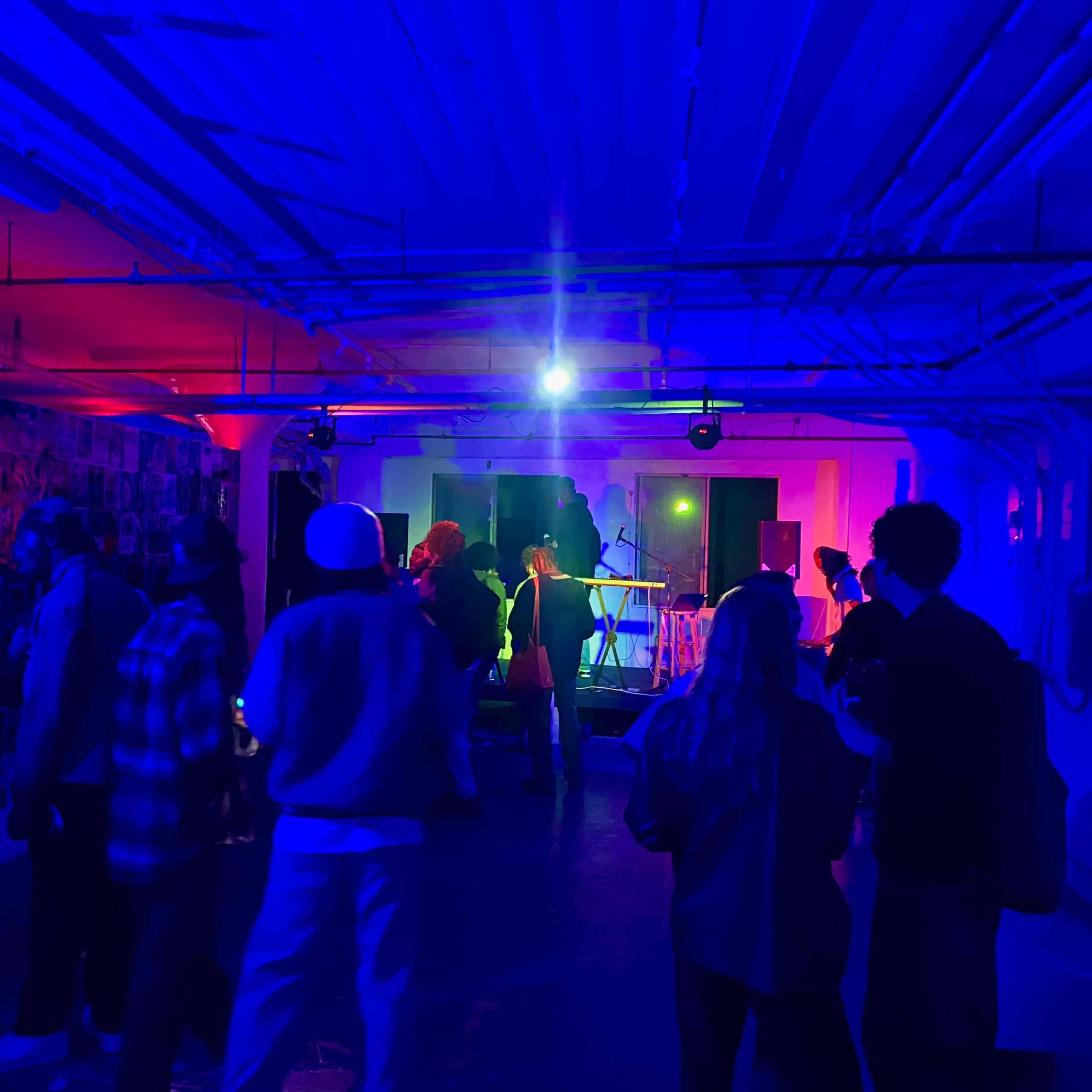 A group of people gathers in a dimly lit room with colorful lights, watching a performance on a small stage.