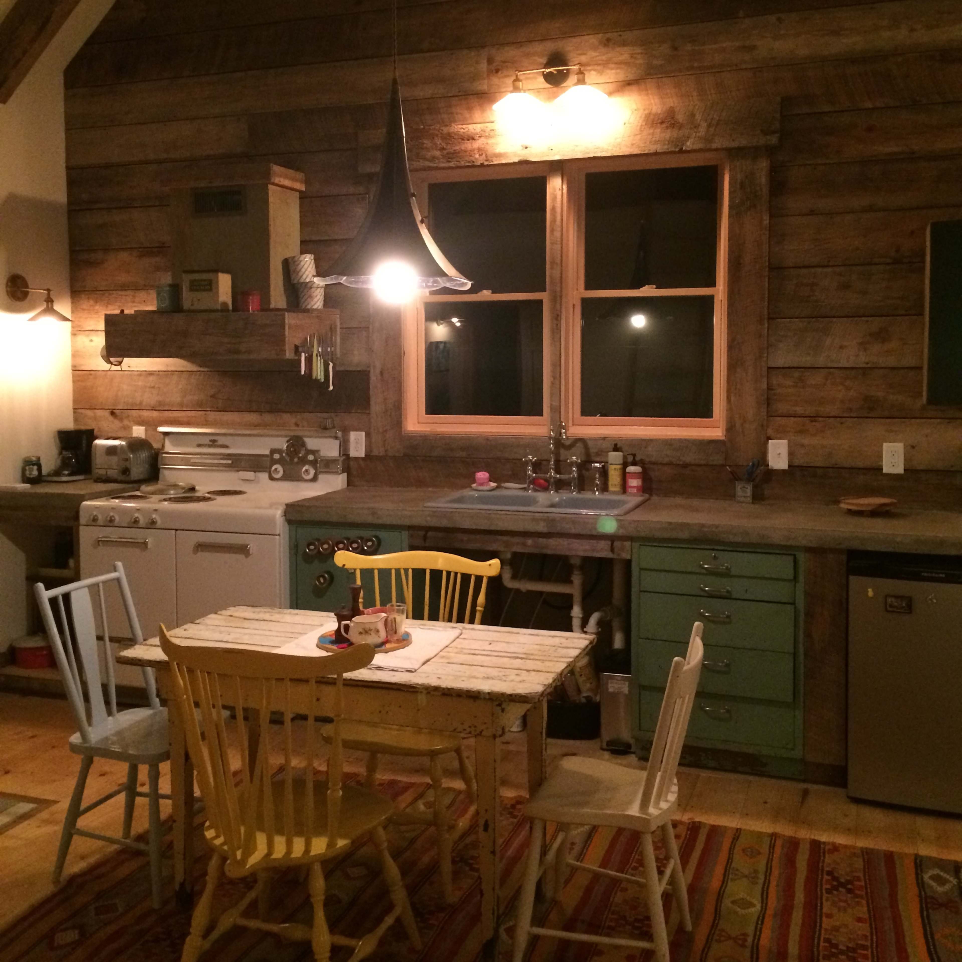A rustic kitchen with wooden walls, a table surrounded by colorful chairs, and a window above the sink illuminated by a pendant light.