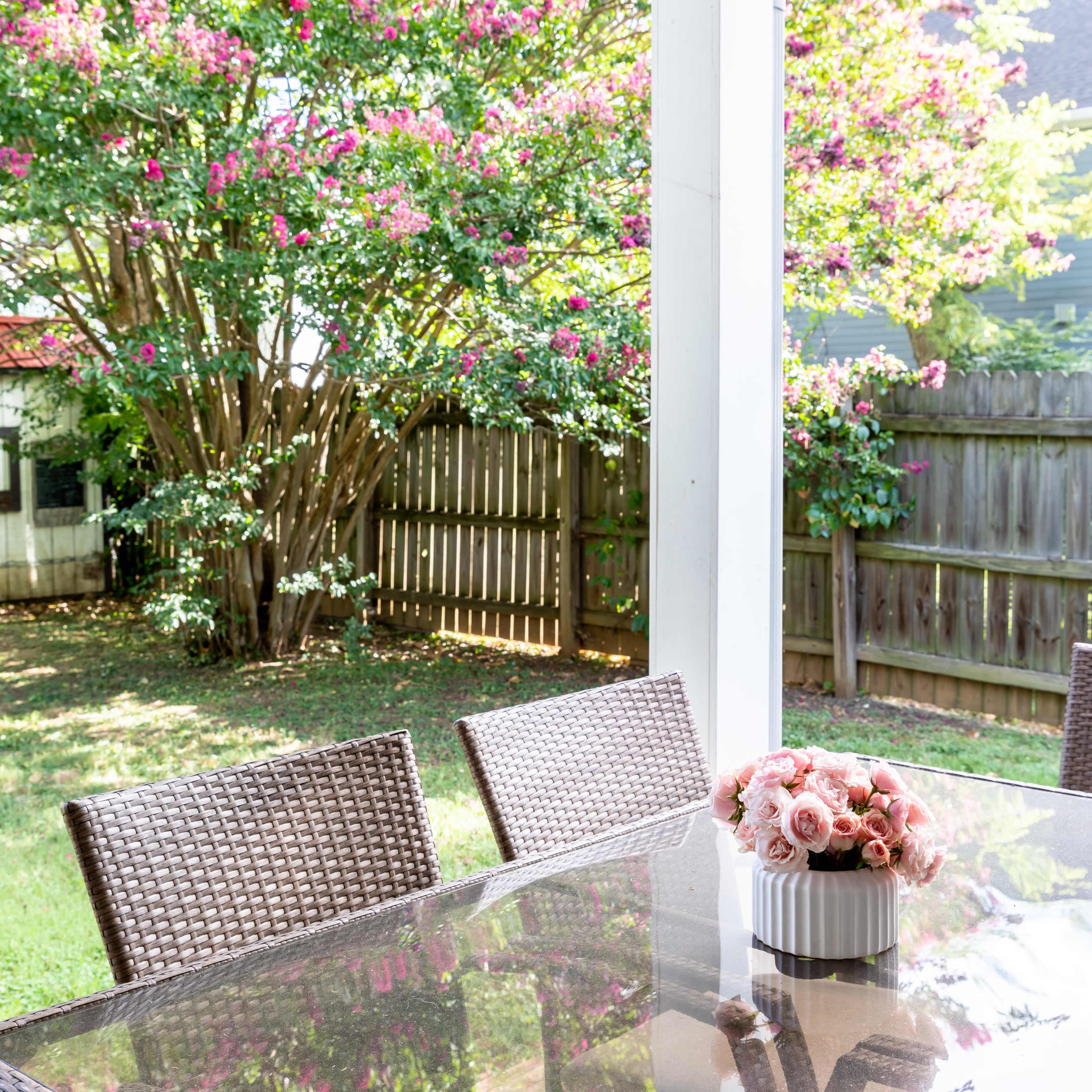A glass-topped table and wicker chairs are set beneath a porch, overlooking a backyard with a flowering tree and wooden fence.