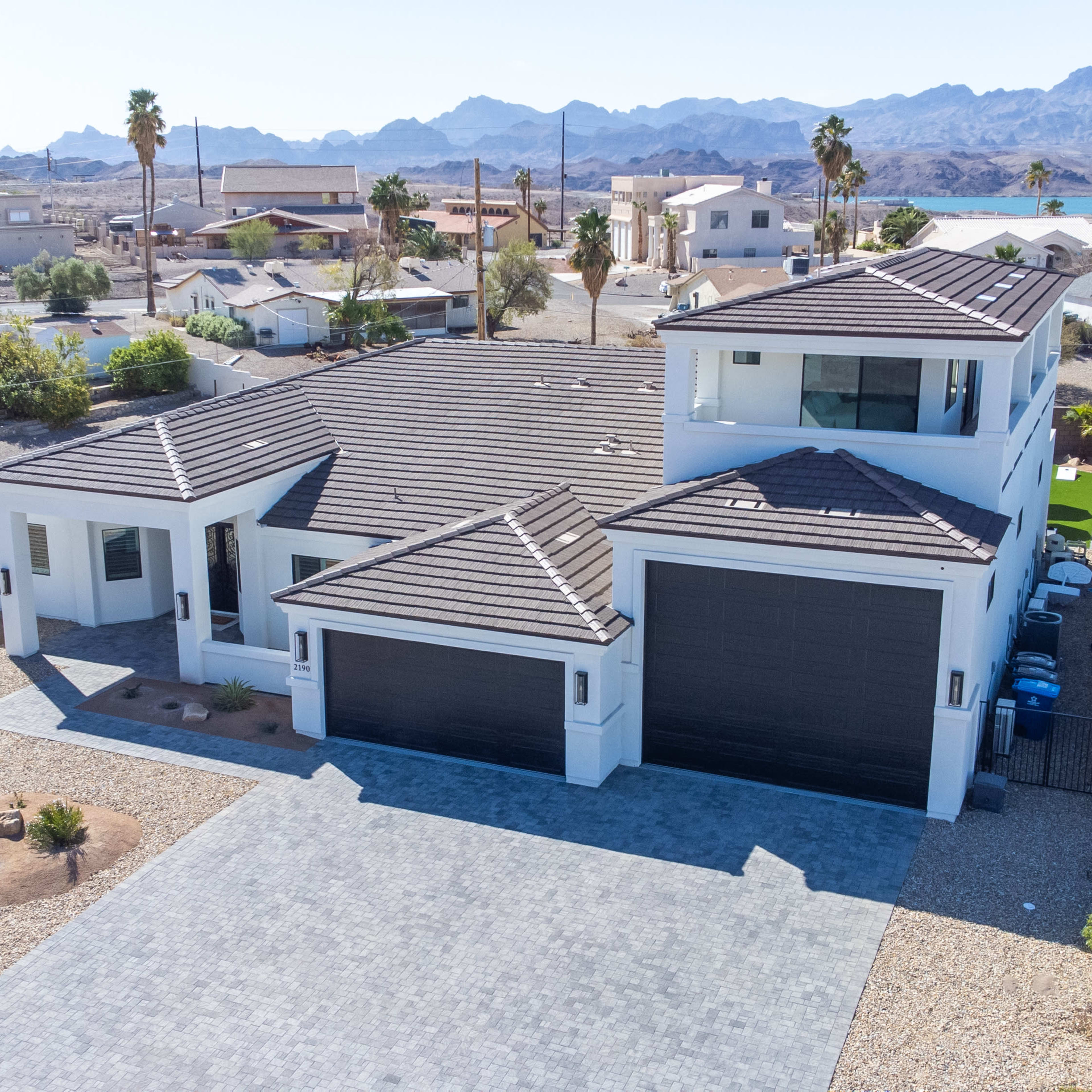 A modern white house with a dark tiled roof and a landscaped yard, situated in a desert area with mountains in the background.
