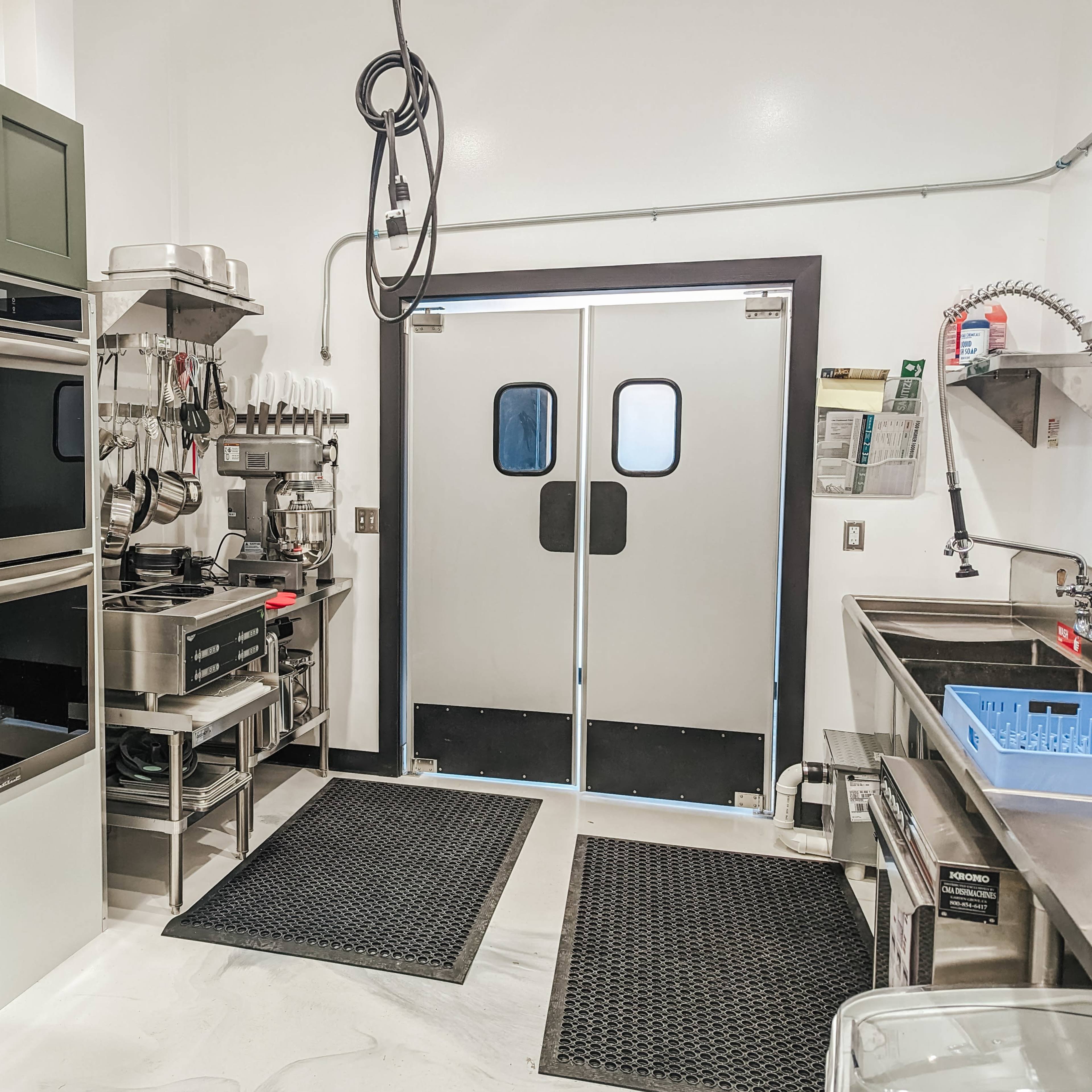 A commercial kitchen area featuring stainless steel appliances, including ovens and a refrigerator, with a double-door entrance and a clean work surface.