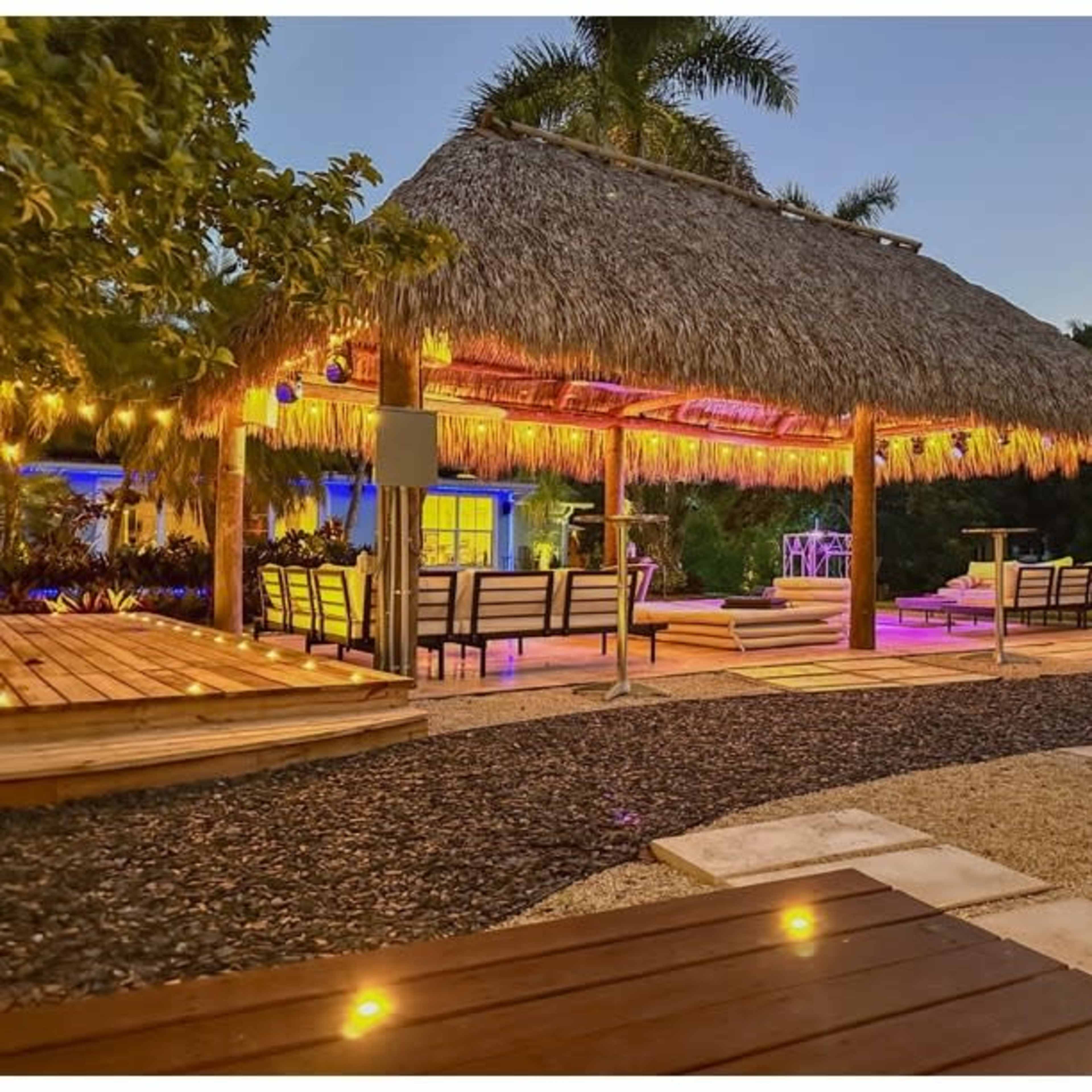 A thatched-roof gazebo with seating and ambient lighting is situated in a landscaped outdoor area.
