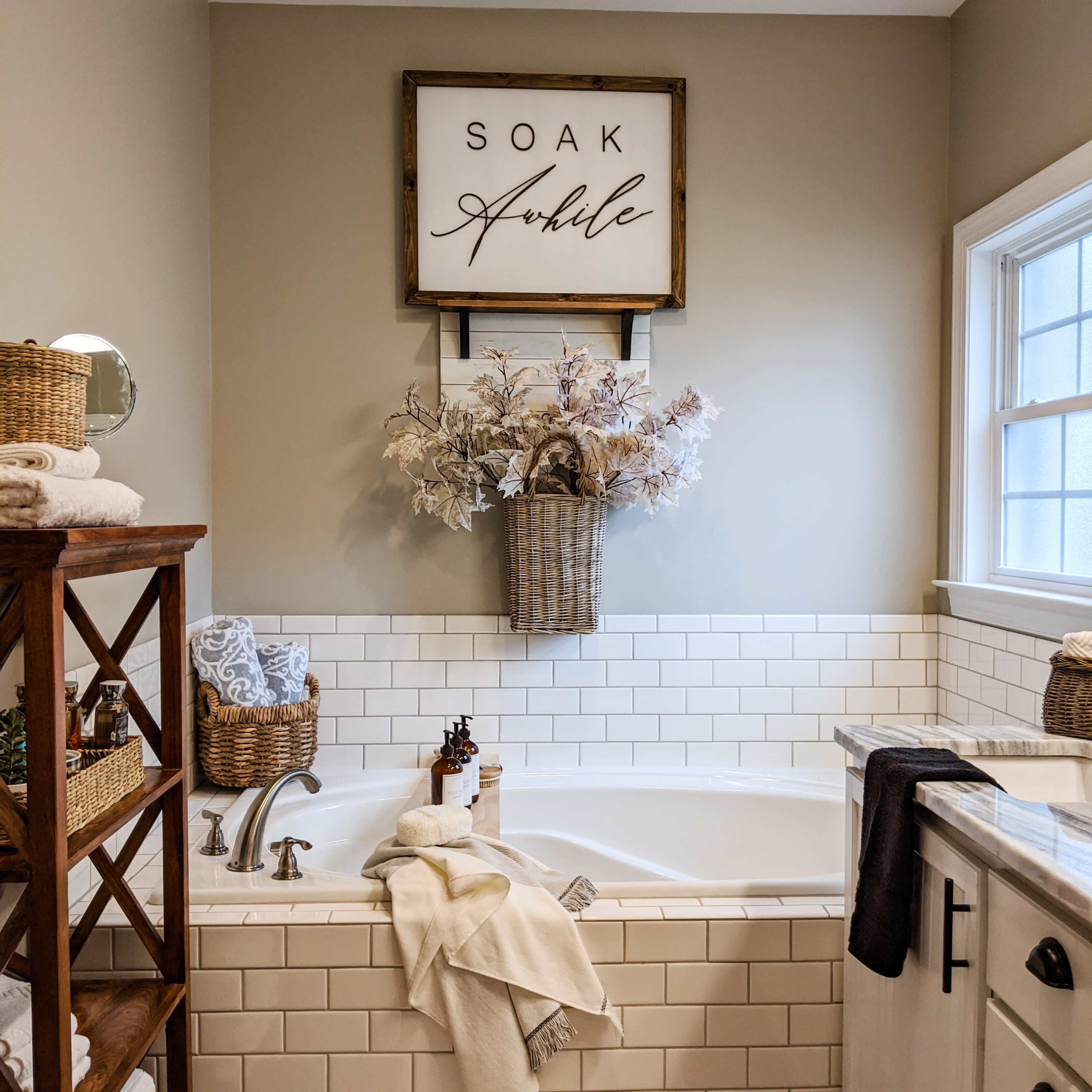 A neatly arranged bathroom features a white bathtub with decorative plants, a wooden shelving unit with towels, and a sign that reads "Soak Awhile."
