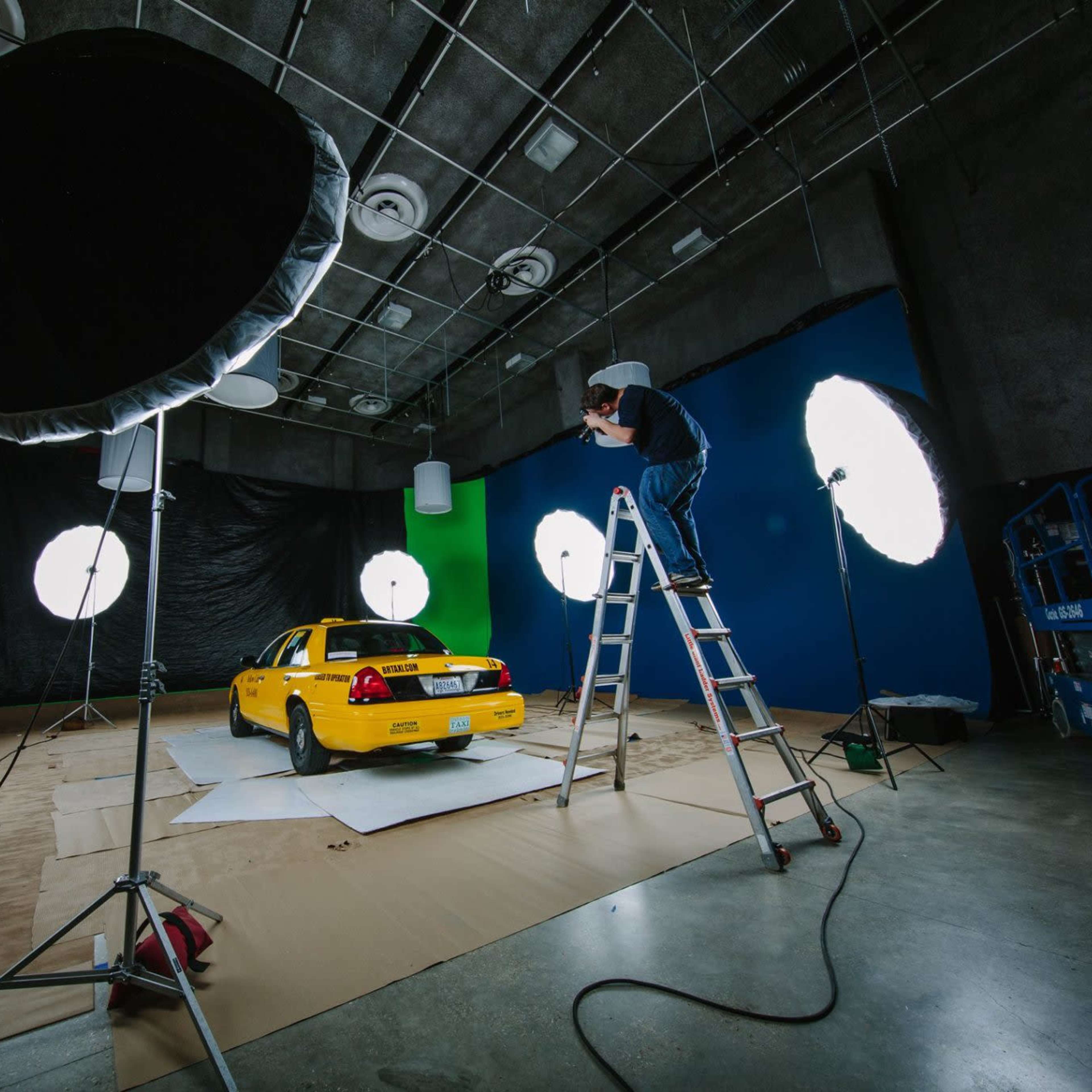 A yellow car is positioned in a studio space with large lighting equipment and a person on a ladder adjusting the lights.