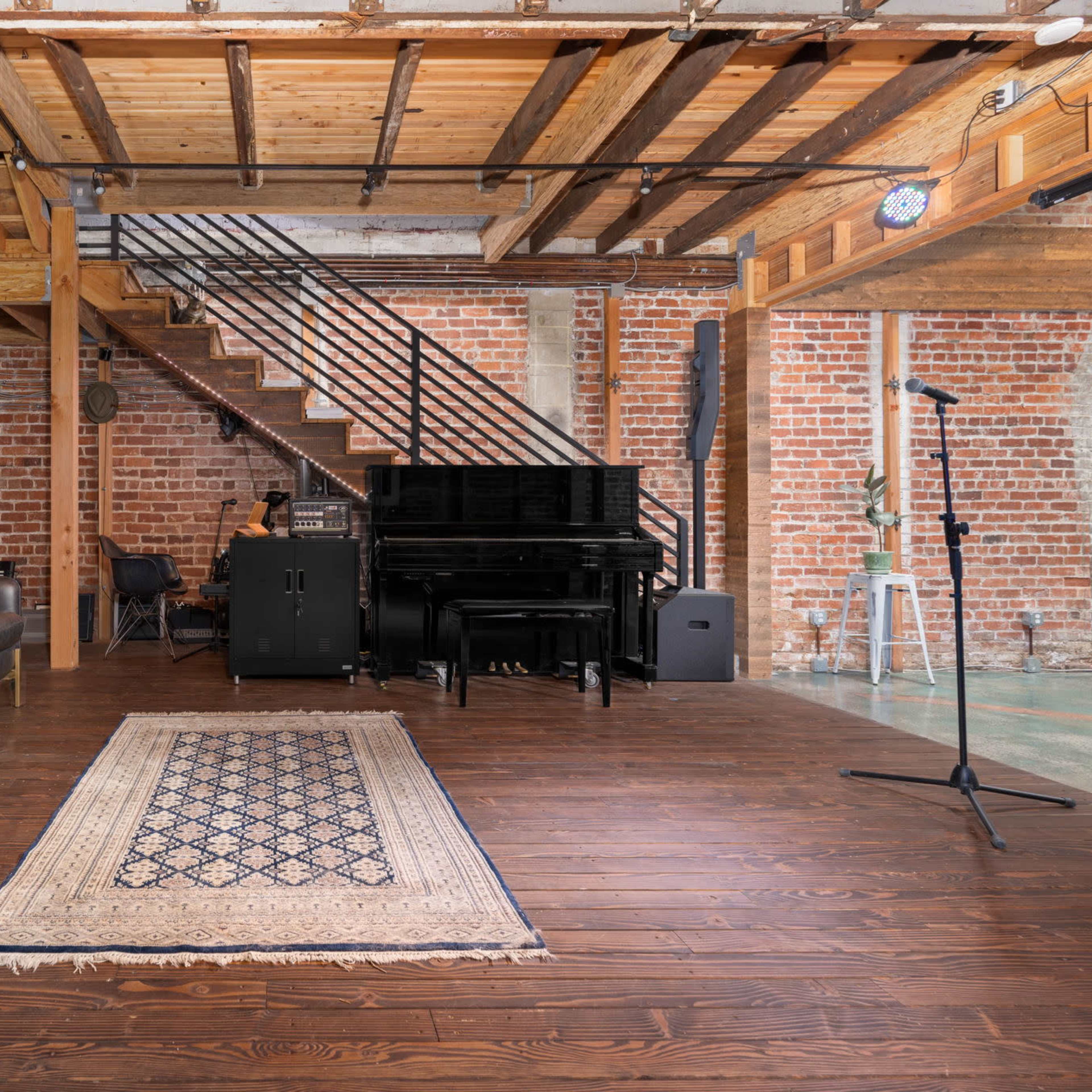 A spacious music studio featuring a leather sofa, a patterned rug, a grand piano, and various guitars mounted on the wall, set against exposed brick and wooden beams.