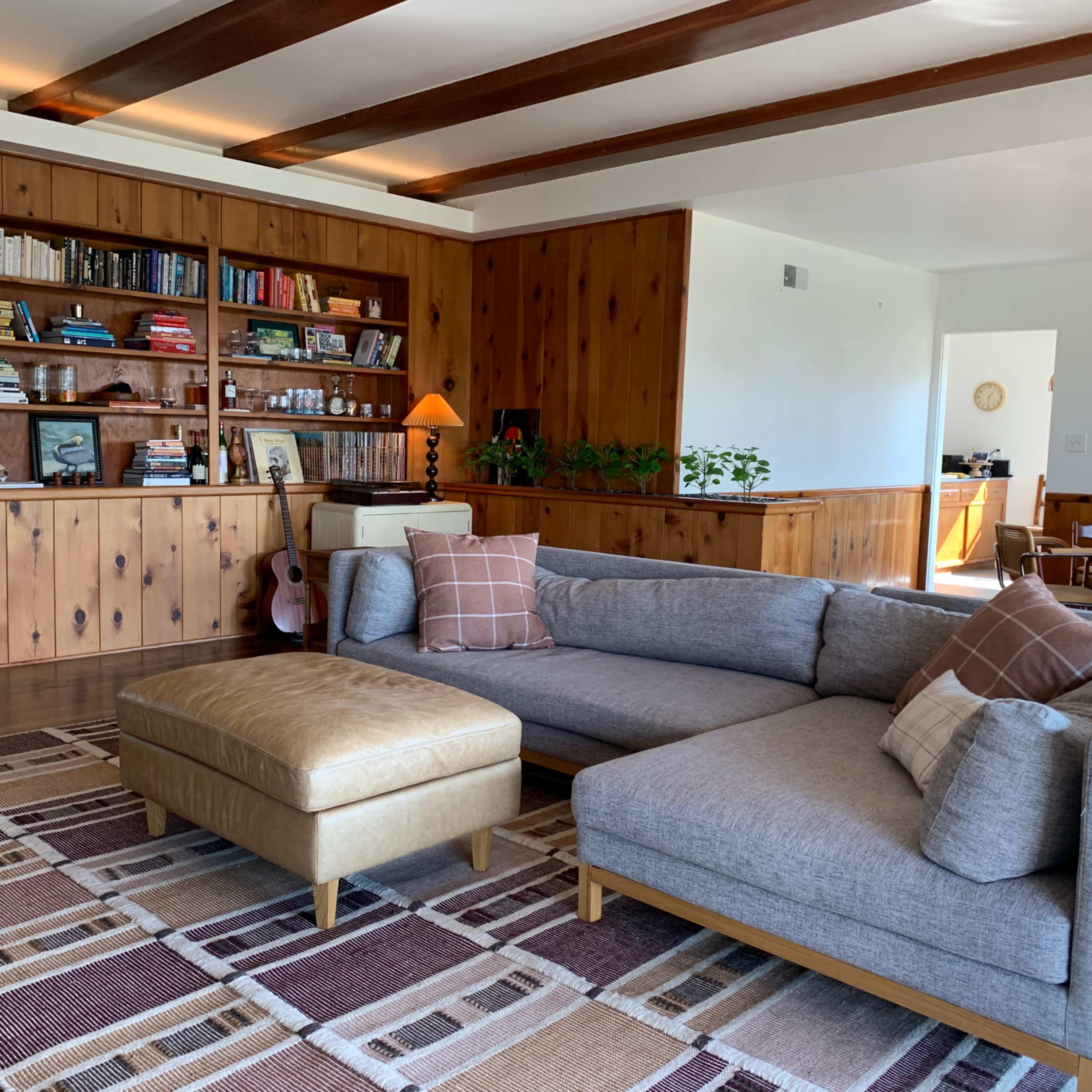A modern living room features a large gray sectional sofa and a leather ottoman, with wooden paneling and built-in shelves filled with books in the background.