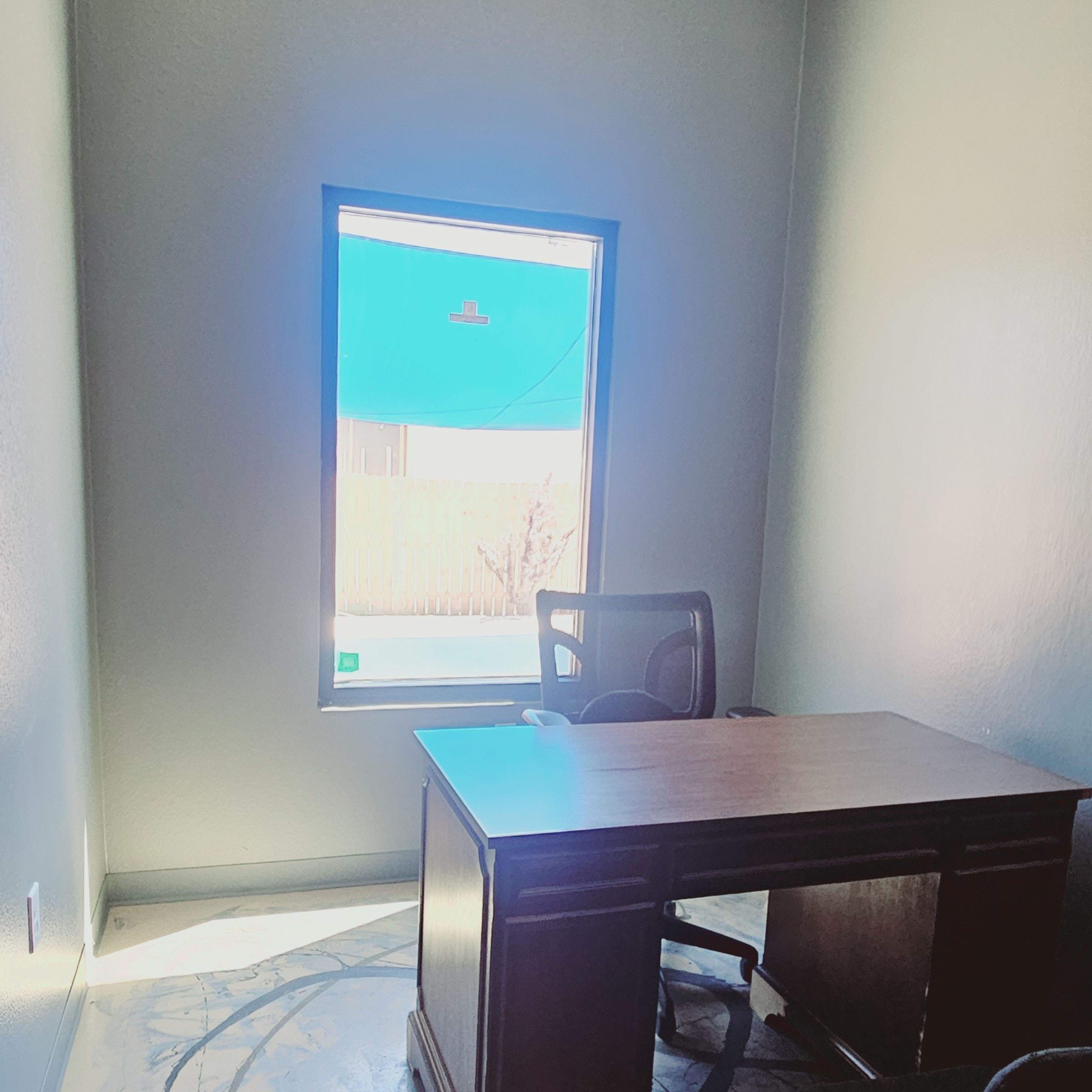 An empty office space features a wooden desk, a black chair, and a large window with natural light illuminating the room.